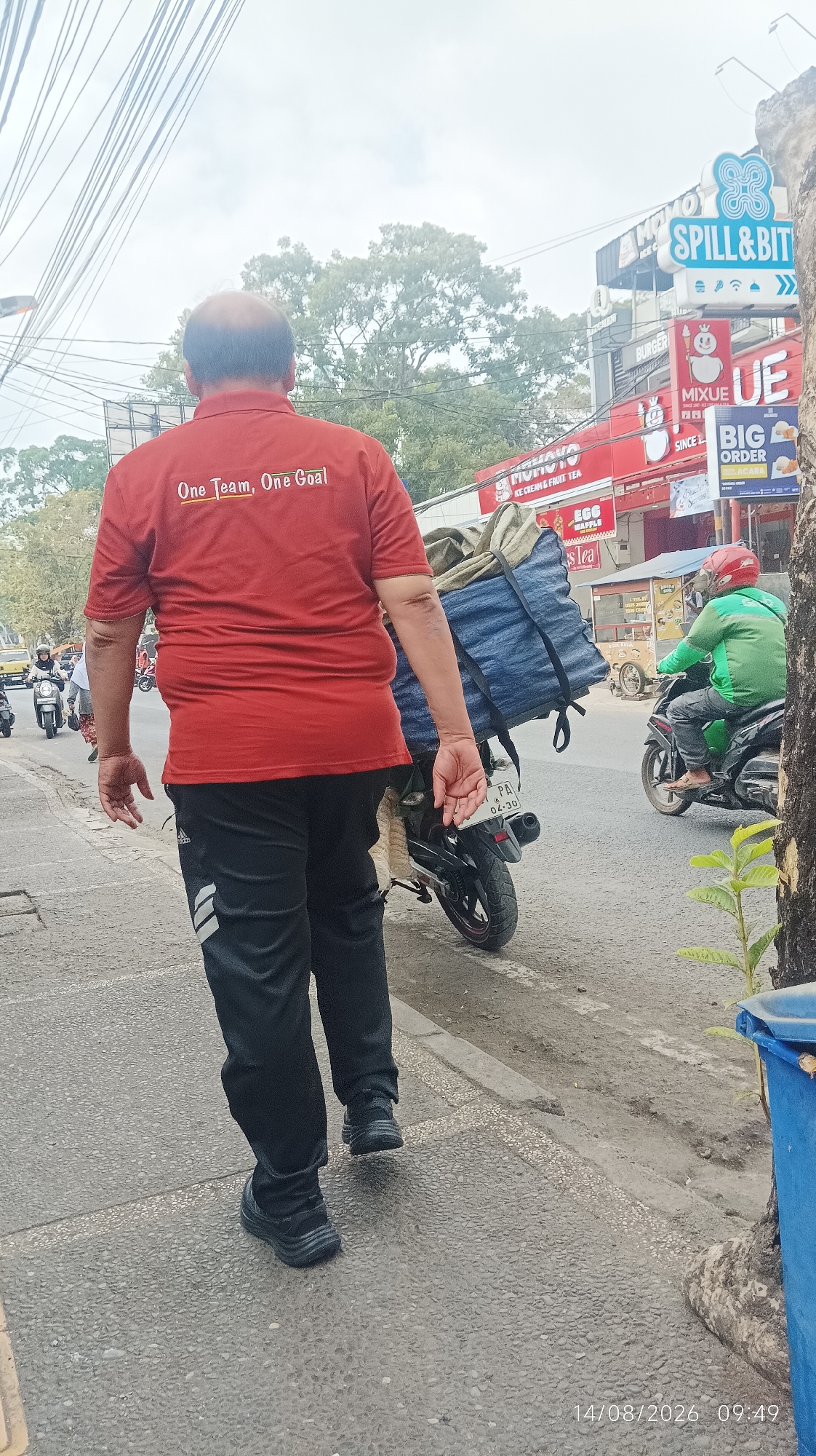 The image shows the rear view of a man in a red t-shirt with the text "One Team, One Goal" printed on the back, walking on a sidewalk. He is wearing black track pants with white stripes and black shoes. A motorcycle with a large blue and green tarp-covered load is parked behind him. In the background, there are several buildings with signs, including "SPILL & BIT," "BURGER," and "BIG ORDER." A person in a green jacket and red helmet is riding a motorcycle on the road. The scene appears to be a street in Tasikmalaya, Indonesia, given the license plate "PA" and the overall urban environment. The timestamp at the bottom right indicates the date and time as 14/08/2026 09:49.