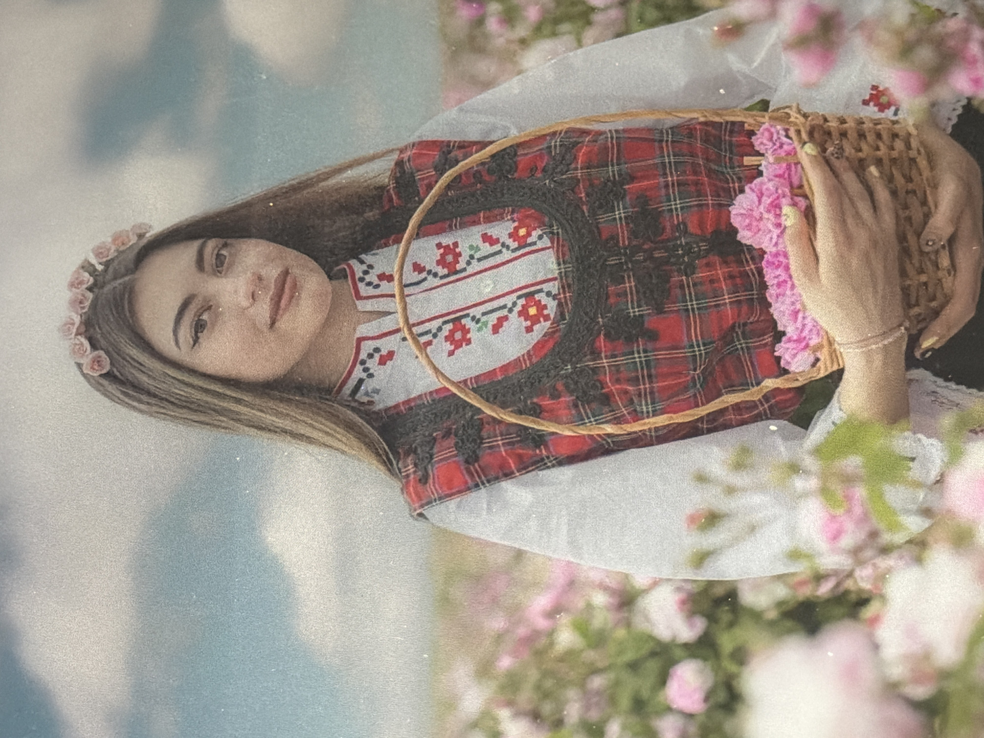 A young woman wearing a traditional red plaid vest over a white embroidered shirt and a flower crown stands in a field of pink roses, holding a basket filled with more pink roses. The sky above is cloudy and blue. The woman has long, dark hair and is looking at the camera. The scene appears to be a rose field, likely for rose oil production, a common practice in regions like Bulgaria.