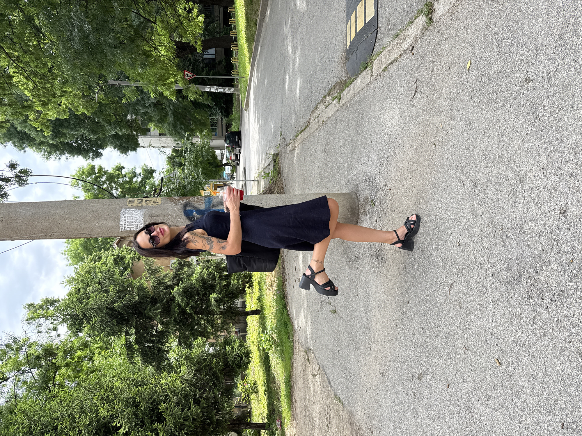 A woman stands on a sidewalk next to a street. She wears sunglasses and a dark dress, and has tattoos on her left arm. She is holding a cup with a reddish liquid. She is wearing black sandals with a thick sole and heel. She is leaning against a concrete post. Trees and other greenery are visible in the background. In the far background, across the street, are parked cars, a street sign, and buildings. The location is Pleven, Bulgaria.