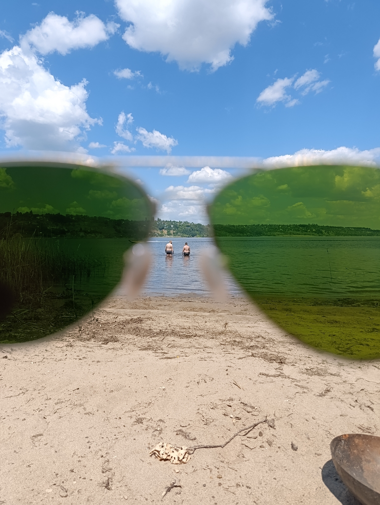 The image depicts a sunny day at a sandy beach, with a body of water in the background. Two individuals are standing in the water, facing away from the camera. The foreground is dominated by sand and scattered debris, with a brown, bowl-like object on the right side of the frame. The scene is viewed through what appears to be sunglasses, casting a green tint over much of the image. The background shows a tree-lined shore and a blue sky with scattered clouds. The setting is likely a riverbank or lake. While no specific landmarks are visible to pinpoint the exact location in Saratov, Russia, it is a generic riverside scene characteristic of areas around the city.