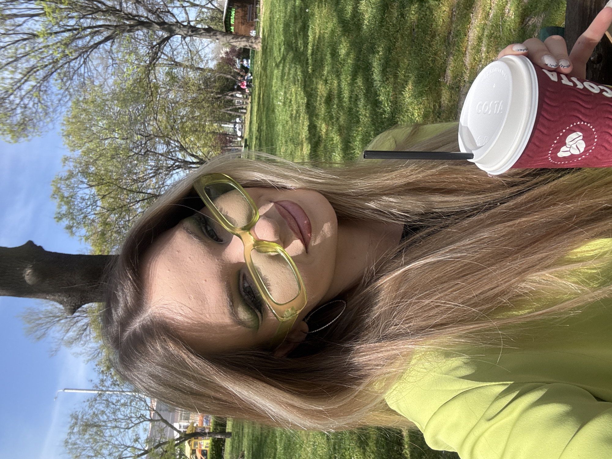 A close-up, brightly lit outdoor portrait features a person with long, blonde hair, wearing clear or light yellow-framed glasses and a lime green top. The person's face is partially visible, showing green eye makeup and a relaxed expression with the tongue slightly extended. One hand, with manicured nails featuring a glittery design, holds a red Costa coffee cup with a white lid and a black straw.
The setting appears to be a park or a large grassy area under a bright blue sky. Numerous trees, some with early spring foliage and others with bare branches, are visible in the background, casting shadows on the vibrant green grass. The overall impression is a sunny day, likely in spring or early summer, in a public green space. The person seems to be enjoying a relaxed moment outdoors.
Notable details include the "COSTA" branding clearly visible on the coffee cup. In the background, a partially obscured sign shows the text "JA PARODC". The location context is Plovdiv, Bulgaria.