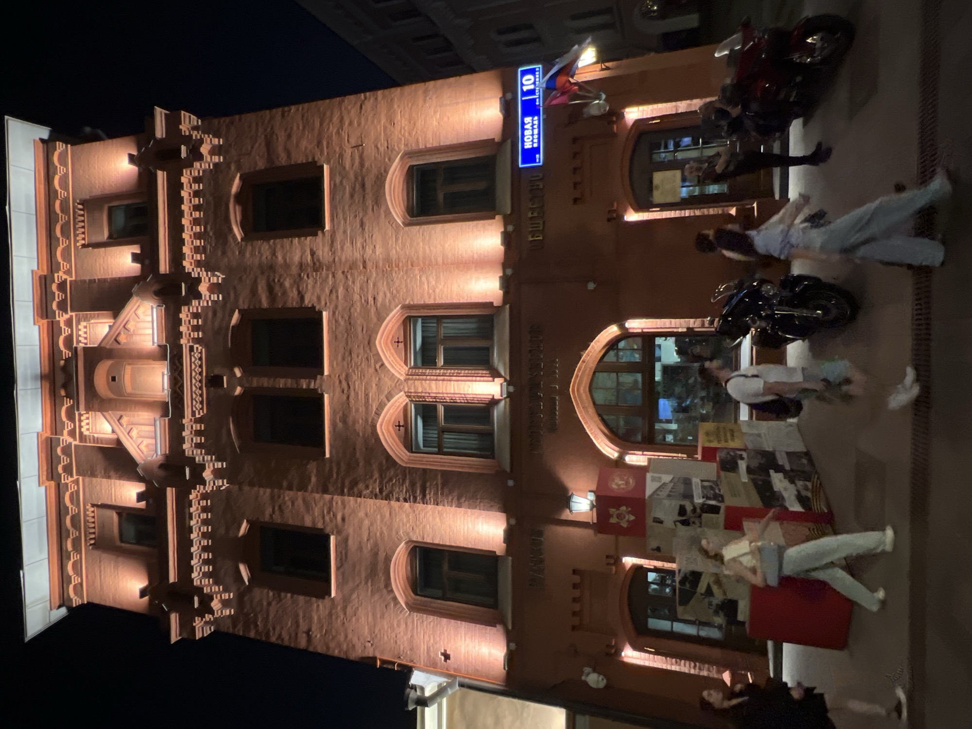 In an outdoor night scene in an unknown city, a tall, red brick building is illuminated with warm-toned lights. The building features multiple floors with arched windows and ornate architectural details along the roofline. A blue street sign with Cyrillic lettering and the number "10" is mounted on the right side of the building, alongside two flags. In front of the building, several people are walking on the sidewalk. To the left, two women are walking. In the center, a woman in light-colored pants and a cream-colored top is walking and carrying a light blue bag. To her right, a man in a white t-shirt and shorts is walking. Further to the right, two motorcycles are parked, and a man is standing near the entrance of the building. A display of historical images is set up on the left side of the sidewalk, near the entrance. The ground appears to be paved.