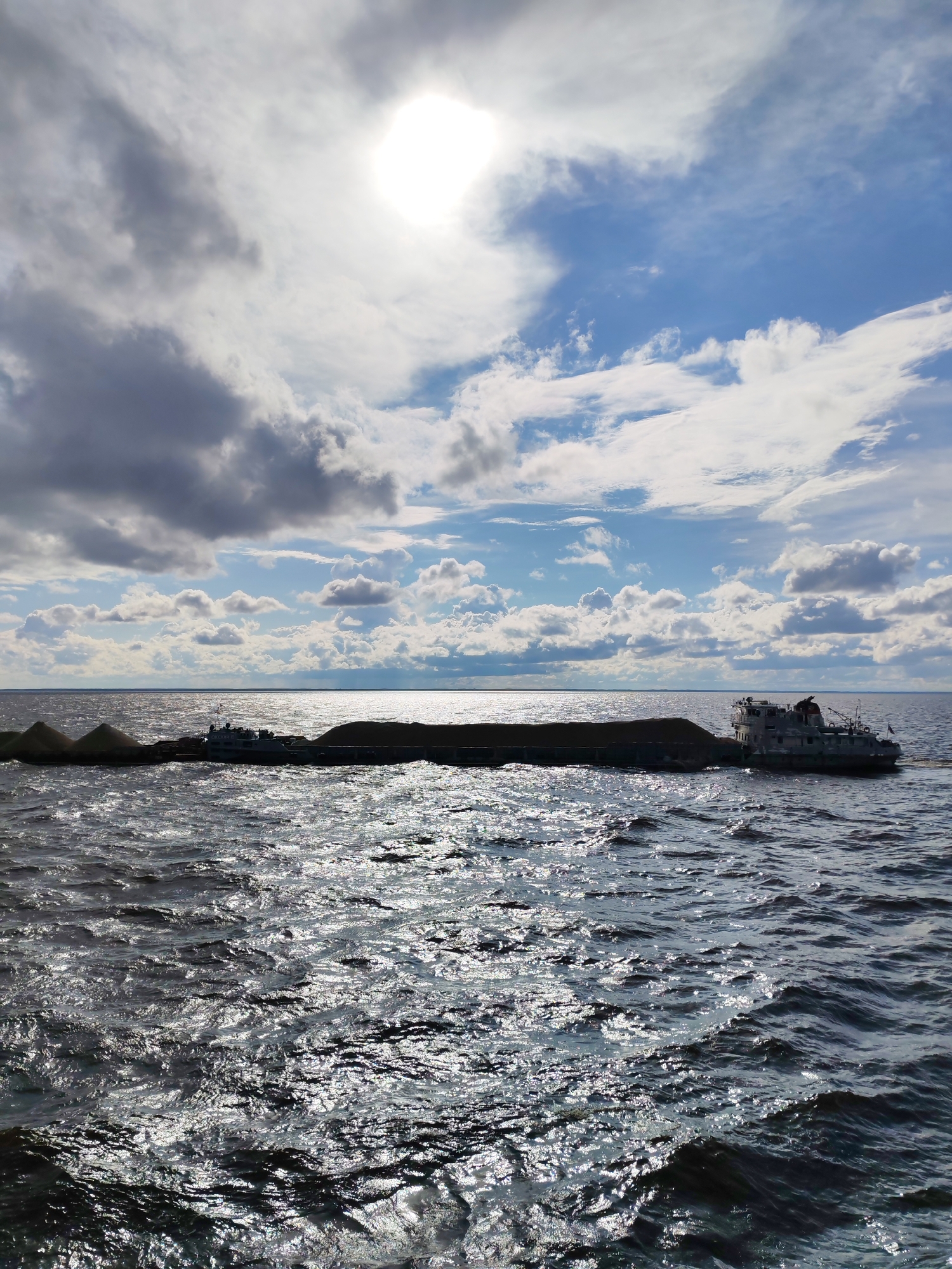 The image shows a barge loaded with dirt or sand being towed by a tugboat on a body of water. The sky is partially cloudy with the sun shining through, creating reflections on the water. The water is choppy, indicating some wind. The barge is long and rectangular, with the cargo piled high in the center. The tugboat is at the stern of the barge, pulling it forward. The scene appears to be a commercial or industrial operation. The location is identified as Prislon', Russia.