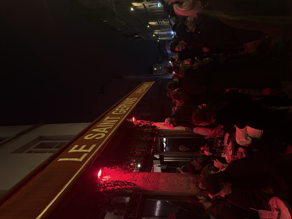 The image depicts a crowded nighttime street scene in Paris, France, specifically in front of the establishment identified as "LE SAINT GERVAIS RESTAURANT." The red-hued illumination originates from lights affixed under the restaurant's dark red awning, which prominently displays the establishment's name in gold lettering.
In the foreground and to the left, several individuals are seated at small, round outdoor bistro tables, engaging in conversation. One table shows a glass containing a clear liquid with ice and a white straw, alongside a pack of cigarettes. Further back and extending across the midground, a dense group of approximately twenty to thirty people stands, appearing to socialize and mill about on the sidewalk. Most individuals are dressed in dark attire, consistent with evening wear. A man wearing a light-colored baseball cap is visible standing near the entrance under the awning. Additional buildings, characteristic of Parisian architecture, line the street in the background, with distant streetlights providing limited ambient illumination further down the block under a dark night sky. The overall scene indicates a busy, social atmosphere at an urban dining or drinking establishment.