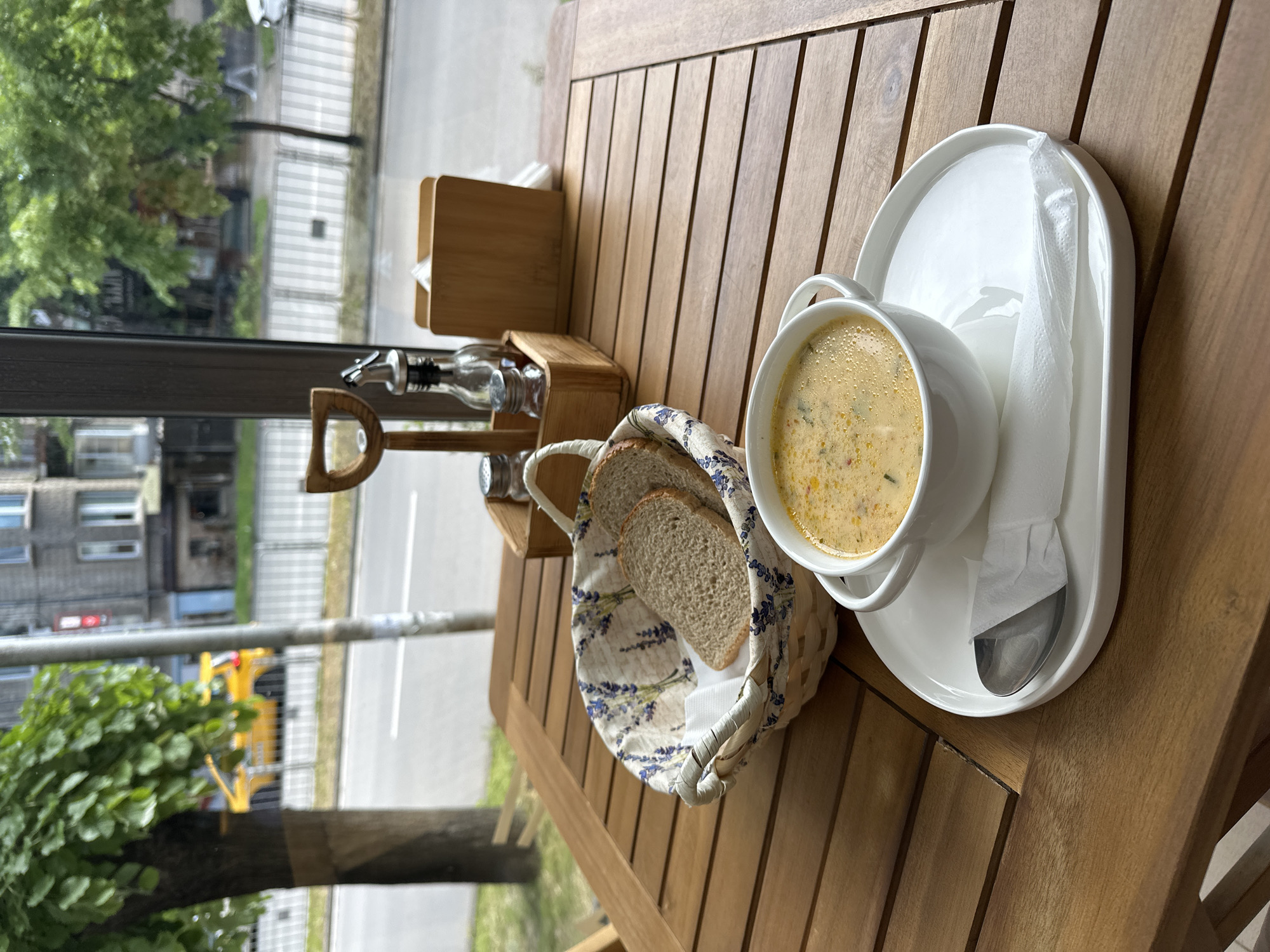A close-up, high-angle shot depicts a table setting, likely outdoors in Varna, Bulgaria, given the visible urban surroundings. The table is made of light brown wood slats. A bowl of creamy soup sits on a white saucer with a white oblong plate behind it. A spoon rests next to the bowl. Adjacent to the soup is a bread basket, lined with a floral-patterned cloth and containing slices of brown bread. A wooden box holding napkins is placed further back on the table, next to a bottle of vinegar and oil. In the background, a window overlooks a street with trees, buildings, and parked cars. A yellow object, possibly a vehicle, is visible amidst the greenery. The focus is sharp on the food and table setting, with the background slightly blurred.