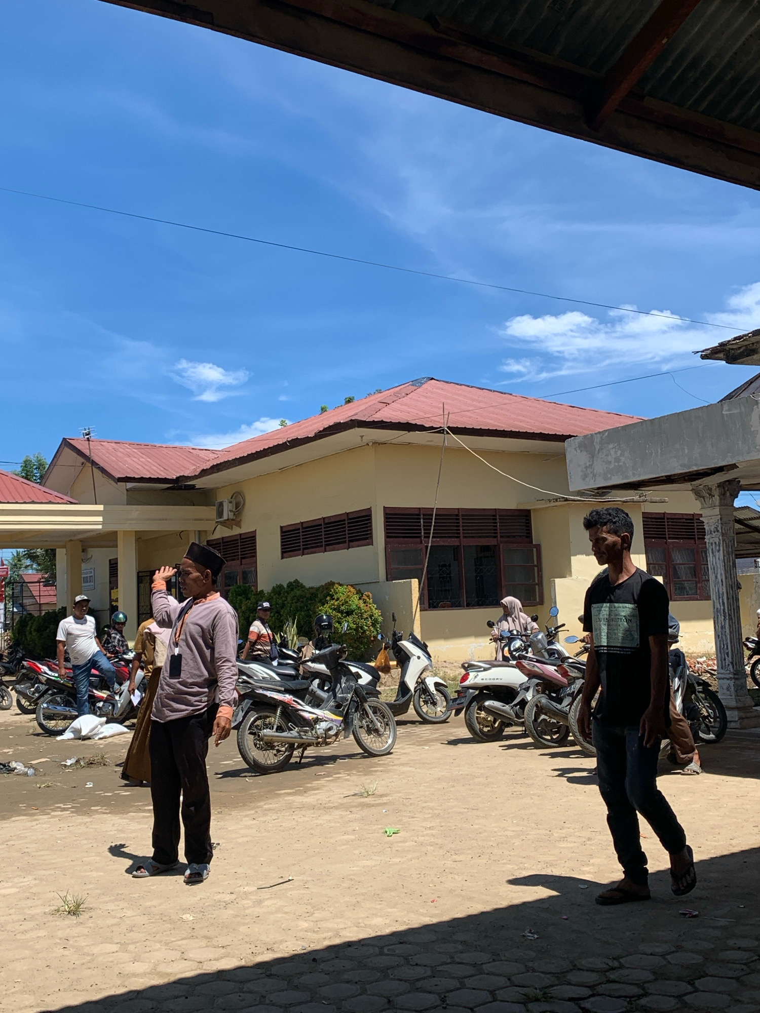 The image depicts an outdoor scene on a sunny day in Pantegurah, Indonesia, near a pale yellow building with a red tiled roof. Several motorcycles are parked in the foreground and midground, on a dirt and gravel surface that transitions to a paved hexagonal stone pattern in the lower right.
In the foreground, a man wearing a dark t-shirt with a graphic print and dark pants walks towards the right, casting a long shadow. To his left, another man in a light brown long-sleeved shirt and dark pants, wearing a black cap, raises his right hand as if waving or shielding his eyes. He has a dark tag attached to his shirt.
Behind the man in the cap, several other individuals and motorcycles are visible. One man in a white t-shirt and blue jeans stands near a red motorcycle. Another person, possibly wearing a headscarf, is seated on a white scooter further back. A person in a striped shirt is also visible near a motorcycle.
The building in the background has multiple windows with dark frames and what appear to be louvers. Bushes and trees are present around the building. The sky is blue with scattered white clouds. A thin wire stretches across the sky. The overall impression is a casual, community gathering or waiting area, common in many Indonesian towns.