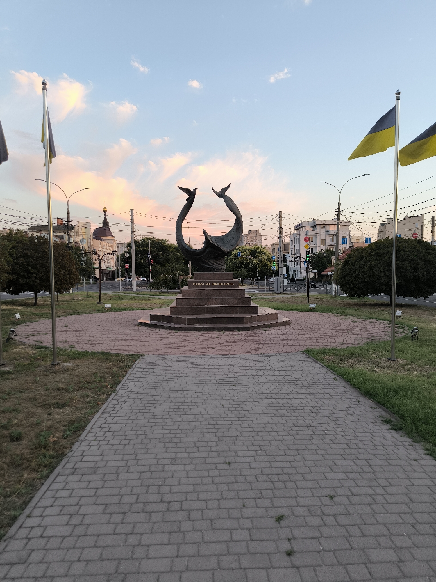 In Mykolaiv, Ukraine, a monument featuring two bronze doves in flight stands prominently in a public square at dusk. The monument is centered on a tiered, stone pedestal. Two Ukrainian flags fly on flagpoles to the left and right of the monument. A paved walkway leads towards the monument, bordered by grassy areas and trees. In the background, buildings, streetlights, traffic signals, and a distant church dome are visible. The sky is painted with hues of pink and orange from the setting sun, with scattered clouds.