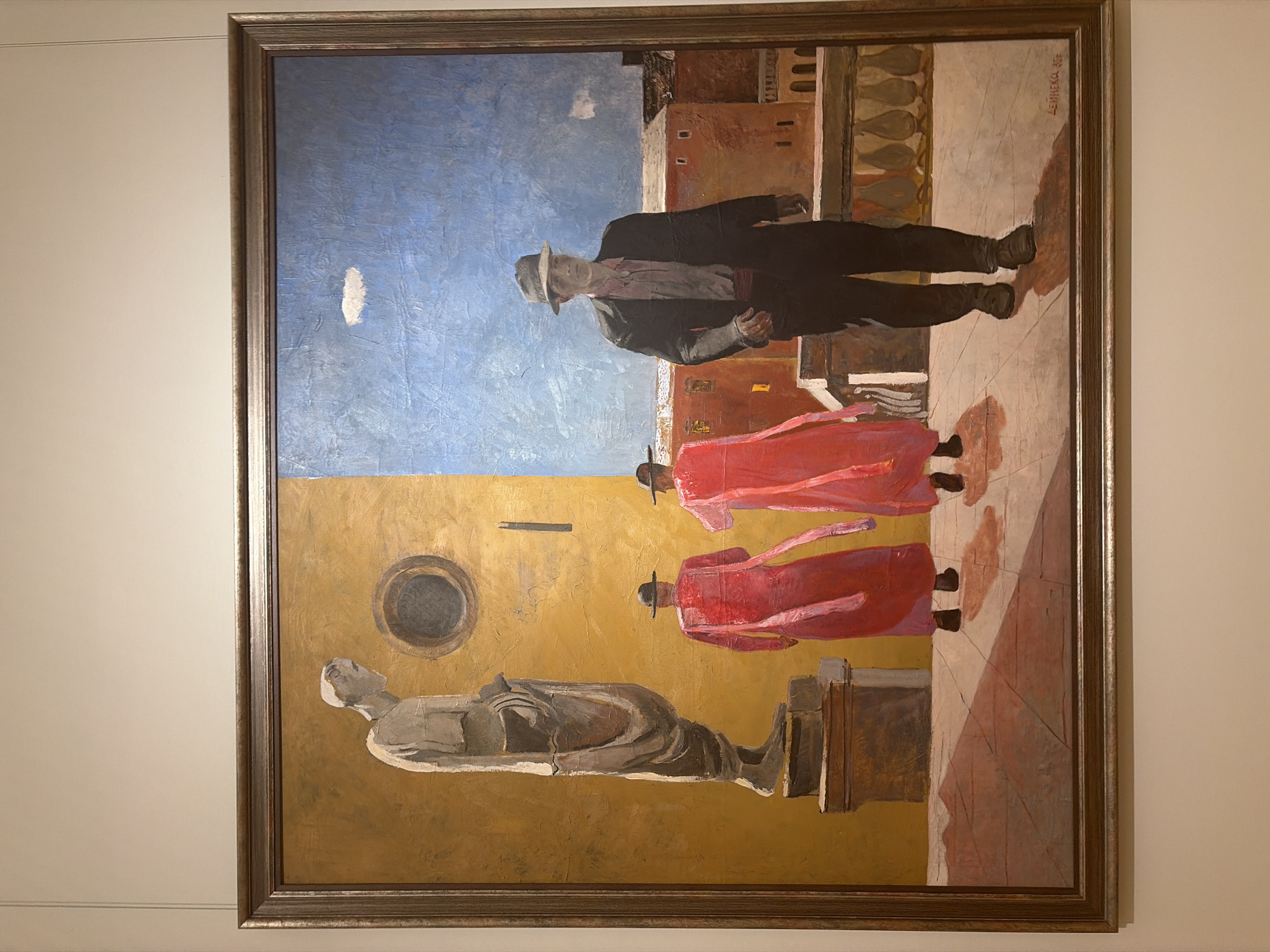 This is a painting, likely oil on canvas, framed in a simple metallic-toned frame. The painting depicts a surreal scene with elements of surrealism and possibly some cubist influence. In the upper half, a man in a hat, dark suit, and red sash walks towards the viewer, looking slightly left with a neutral expression. Behind him, two figures in vibrant red robes and hats also walk towards the viewer, their forms stylized and somewhat abstract. They appear to be in a sun-drenched or brightly lit outdoor setting with reddish-brown buildings in the background. The sky is a bold blue with a small white cloud.
The lower half of the painting transitions to a golden-yellow background, featuring a stark, grey, reclining statue or effigy that appears to be made of stone or plaster. This statue lies on a pedestal, possibly representing a historical or mythological figure. To the left of the statue is a dark circular form with a grey interior, perhaps representing a window, portal, or a stylized sun.
The overall impression is dreamlike and enigmatic. The contrast between the distinct figures and the abstract background, along with the juxtaposition of the man and the robed figures with the statue, suggests a narrative or symbolic meaning that is open to interpretation. The lighting suggests daytime, with strong shadows on the right side of the painting, indicating a specific time of day where the sun is casting long shadows. The architecture hints at a Mediterranean or perhaps even a North African setting, though the presence of the statue and the overall mood might also evoke a sense of timelessness or a psychological landscape.
Visible text is present in the upper right corner of the painting, a signature that reads "ЗЕМНЕХОД."