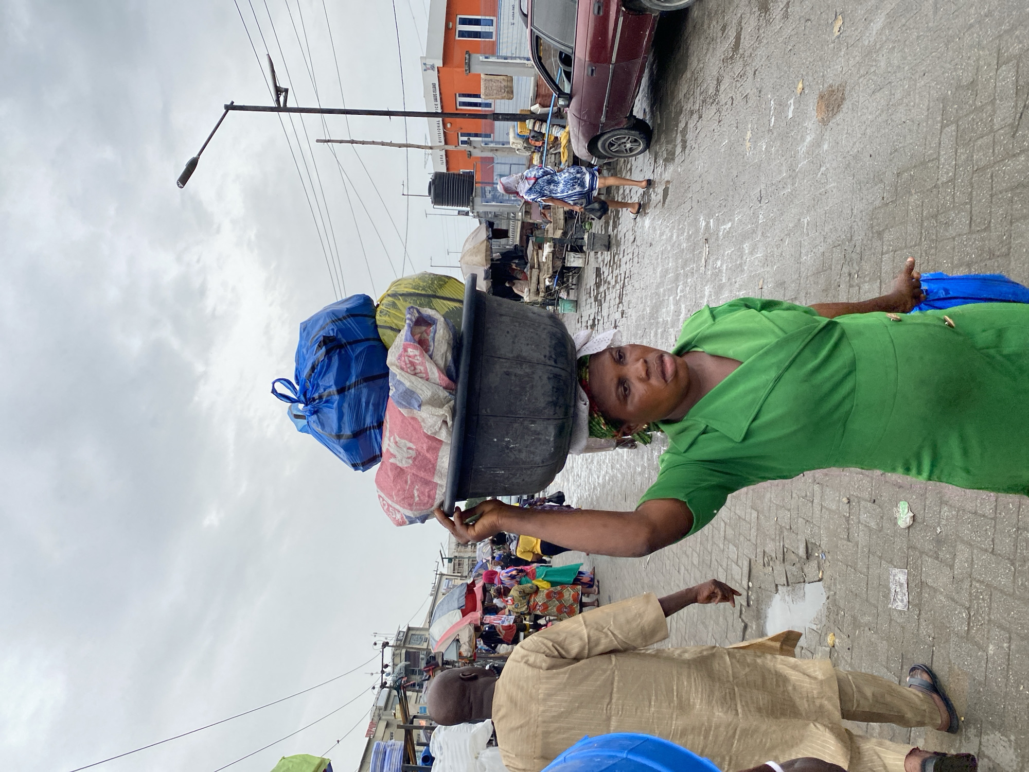 The image depicts an outdoor scene, likely a marketplace or street in Lekki, Nigeria, under an overcast sky. The foreground features a woman balancing a large black tub filled with items on her head, including bundles wrapped in blue, yellow, and patterned fabric. She wears a green dress and a white head covering. A man in a tan outfit stands in the lower part of the frame, with his arm extended, pointing towards something outside the view. In the background, there's a glimpse of a red car, a building painted in orange and white, and a woman in a blue and white dress walking. A street light pole and overhead wires are visible against the cloudy sky. The ground is paved and appears wet, suggesting it might have recently rained. The scene is bustling with activity as implied by the presence of other people and stalls in the background.