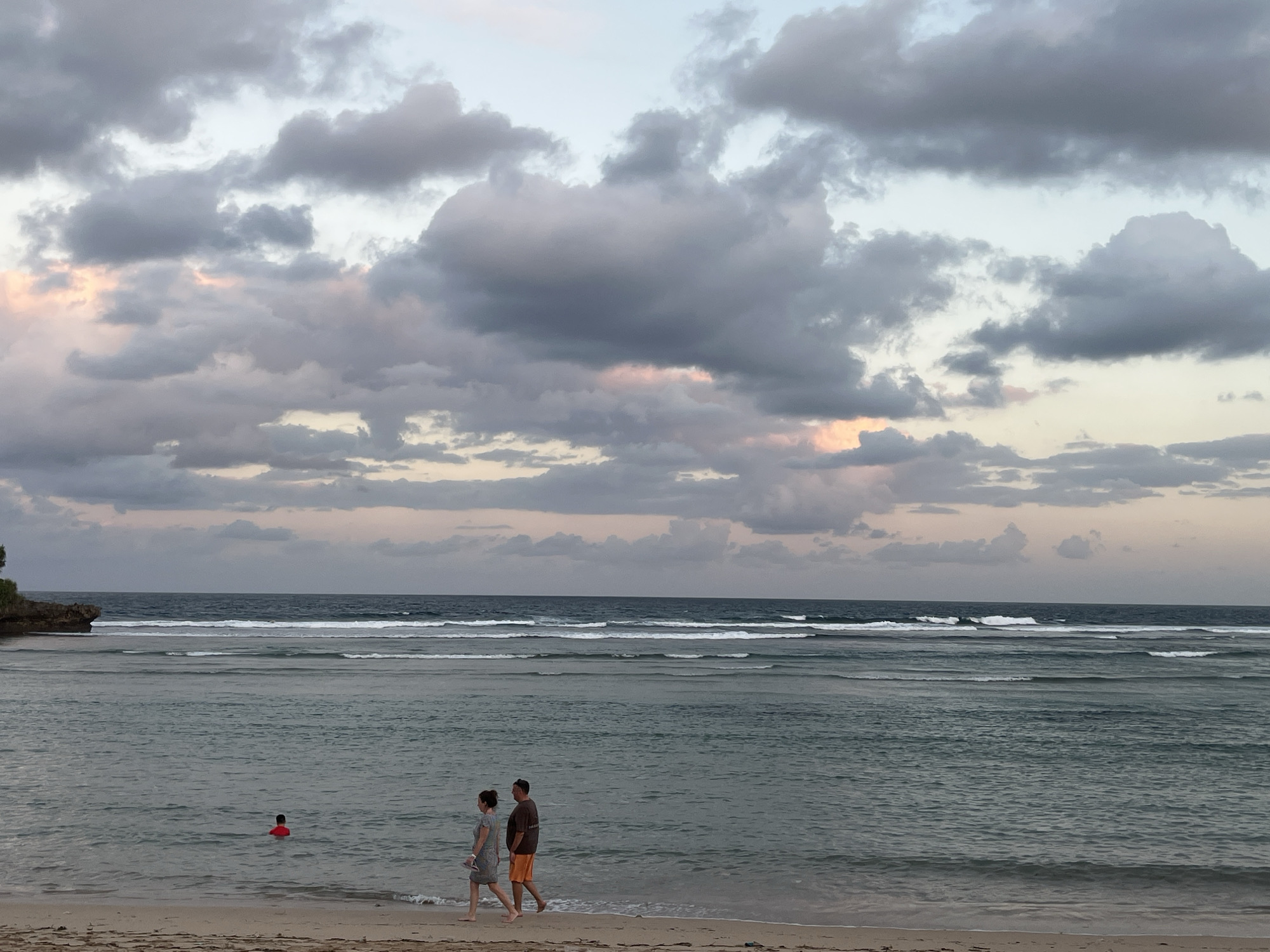 A wide shot captures the shore of a beach in Kuta Selatan, Indonesia, under a cloudy sky. The ocean stretches to the horizon, with gentle waves rolling onto the sandy beach. In the foreground, a couple walks along the water's edge. The woman is wearing a grey dress and the man is in a dark t-shirt and orange shorts. Further out in the shallow water, a person in a red top is visible. The sky is filled with dramatic, textured clouds, some tinged with soft pink and orange from the setting sun. To the far left, a rocky outcrop juts out into the sea, covered in lush green foliage.
