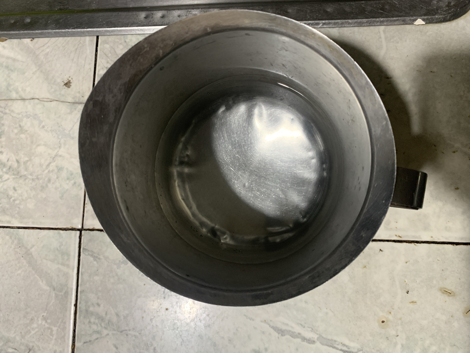 An overhead shot captures a metallic pitcher, possibly stainless steel or aluminum, filled with water, resting on a light-colored tiled floor. The pitcher is round with a slightly flared rim and a handle partially visible on the right side. The water inside is still, reflecting light from an unseen source, creating bright streaks on its surface and the bottom of the pitcher. The interior of the pitcher shows some wear, with visible scratches and indentations.
The floor tiles are square, likely ceramic or porcelain, with a muted, possibly off-white or light grey hue, and feature subtle, darker patterns. Dark grout lines crisscross the floor, indicating wear and possibly some grime. In some areas, particularly noticeable in the cropped images, small specks of dirt or debris are scattered on the tiles and within the grout. A dark, narrow metal surface, possibly the edge of a counter or another appliance, runs along the top of the frame. The overall setting appears to be an indoor space, potentially a kitchen or a utility area in Santa Ana, Venezuela. No people or visible text are present.