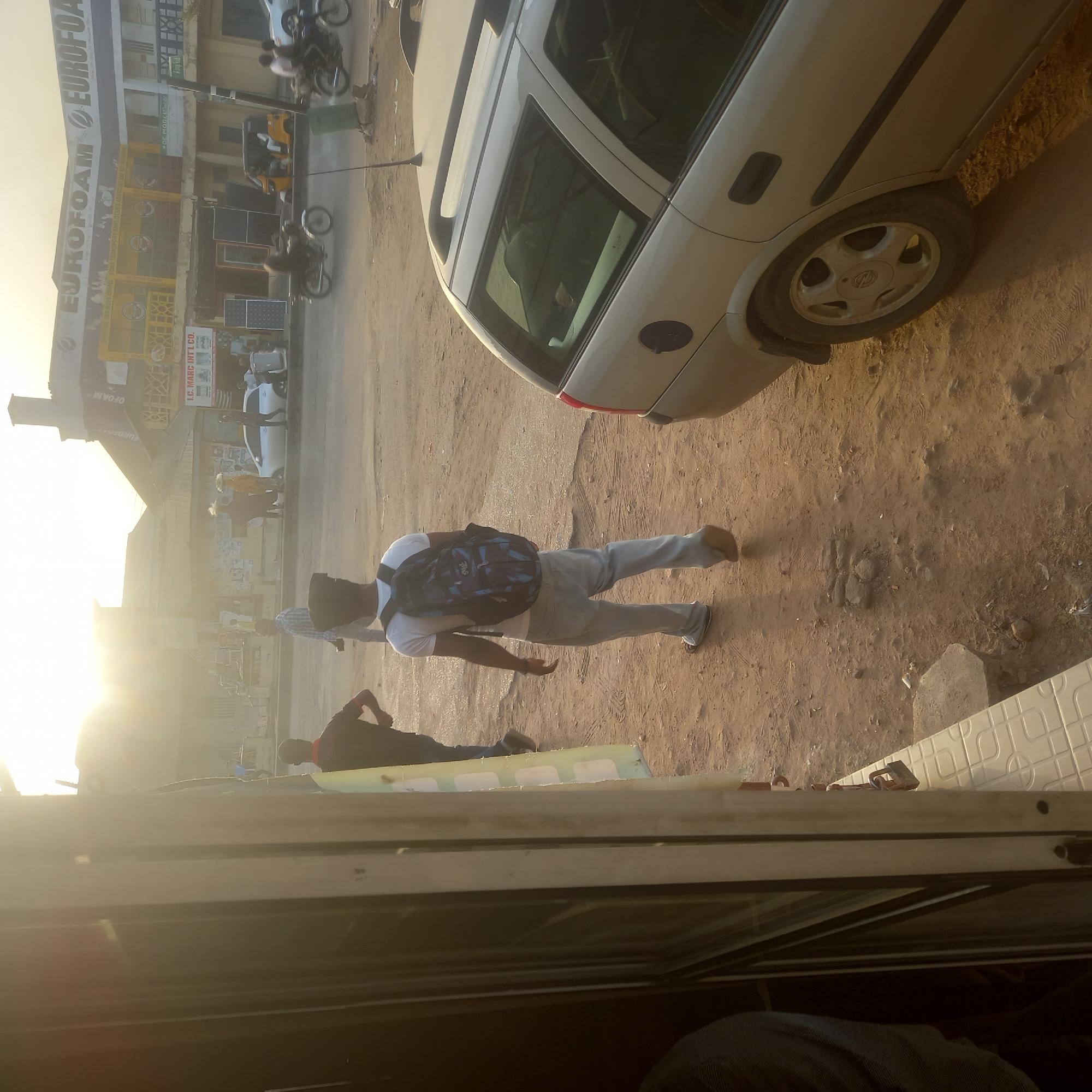 The image appears to be taken in Sardauna, Nigeria. The prominent building to the left has a sign that reads "EUROFOAM". The area is dusty, with a man with a backpack walking near a parked silver car. Another man is visible lying down nearby. In the background, there are motorbikes and other vehicles along the road. The overall scene suggests a daytime setting in a commercial area.