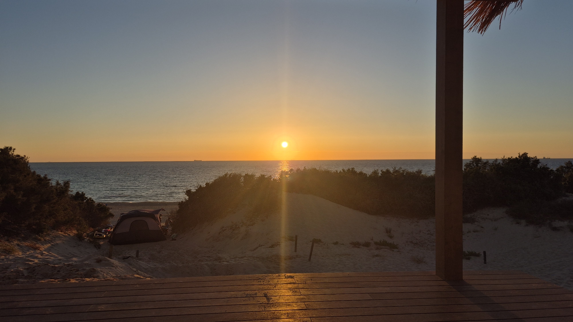 The image captures a sunset over the Mediterranean Sea from an elevated perspective in Ashdod, Israel. The sky transitions from a light blue at the zenith to vibrant orange and yellow hues at the horizon. A bright, circular sun disc is centered just above the horizon, emitting a pronounced vertical sun flare that extends downwards into the water and onto the foreground.
The sea occupies the middle ground, displaying gentle waves and reflecting the sun's light in a shimmering path towards the viewer. Several cargo ships are visible as dark silhouettes on the distant horizon.
In the immediate foreground, a wooden deck with horizontal planks extends across the bottom of the frame, bathed in the orange glow of the setting sun, particularly on the right side. A robust, dark brown wooden pillar or post rises vertically from the right side of the deck, partially obscuring the view, with the edge of a thatched roof visible just above it.
Between the wooden structure and the sea, a landscape of sand dunes with scattered, dark green coastal vegetation is present. Nestled within a sandy depression on the left side, a dome-shaped camping tent, predominantly dark blue and purple, is pitched on the sand, accompanied by some unidentified camping gear. The sand itself shows varied texture and subtle shadows.