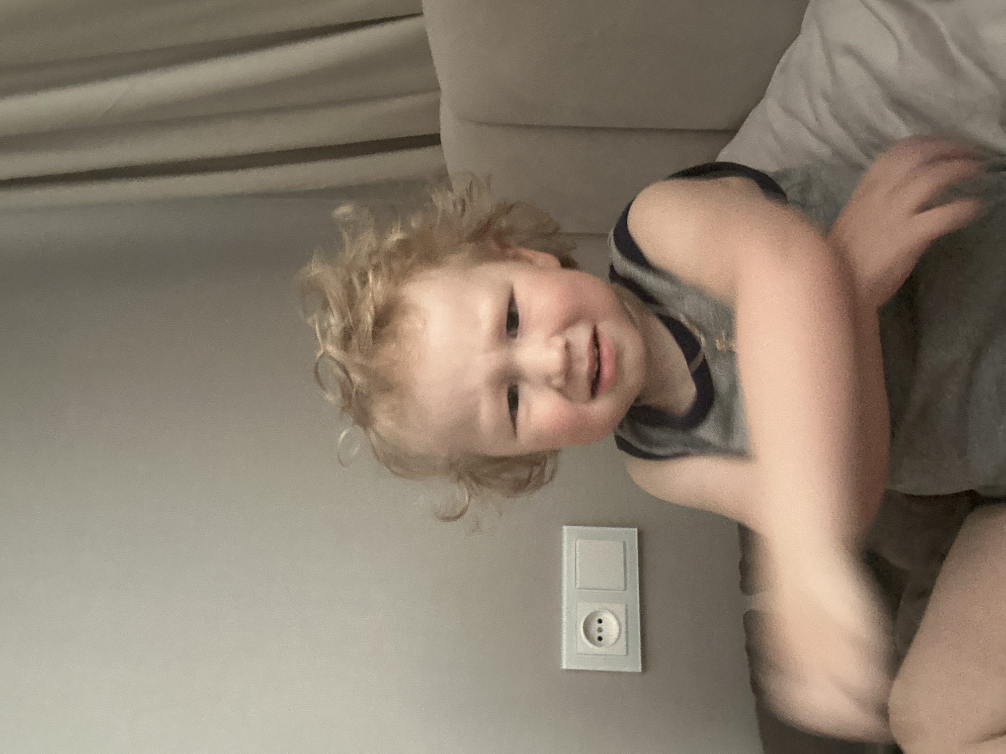 A young, light-skinned child with curly blonde hair lies on a light-colored sofa, wearing a gray sleeveless shirt with a dark blue trim. The child is looking at the camera with a slight grin. The background consists of a gray wall with a European-style electrical outlet. The specific location in Votkinsk, Russia is not discernible from the image.