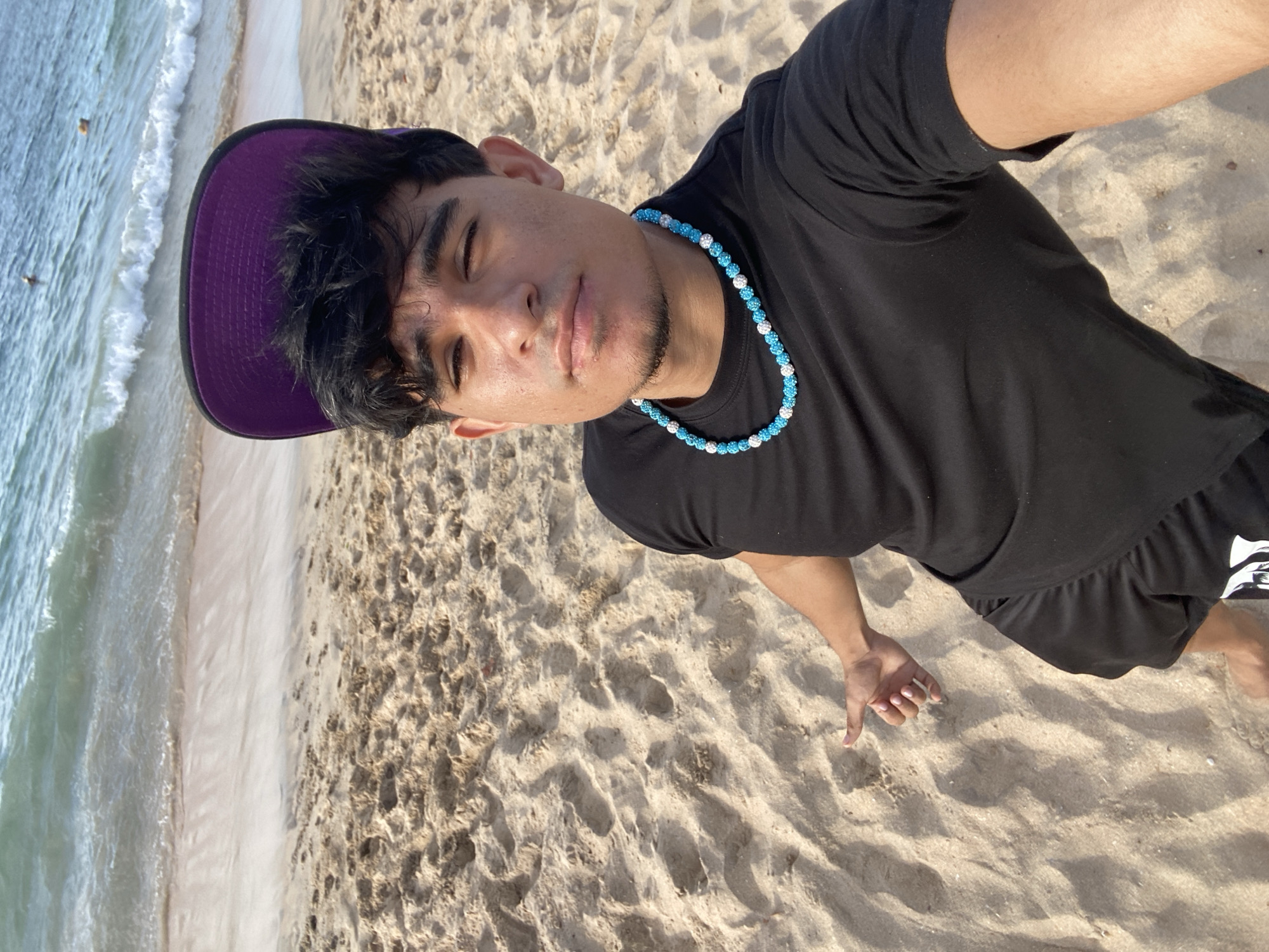 In Altagracia, Venezuela, a young man with dark hair and a light complexion is taking a selfie on a sandy beach. He is wearing a black t-shirt, black shorts, and a purple baseball cap worn backward. A distinctive blue and white beaded necklace adorns his neck. The beach sand is visible in the foreground, with footprints imprinted on its surface. In the background, the ocean waves gently roll onto the shore, and a few people are visible in the water. The man is looking towards the camera with his eyes partially closed, and his left hand is extended outward, fingers partially curled, as if gesturing or posing. The overall scene captures a casual moment on a sunny day at the beach.