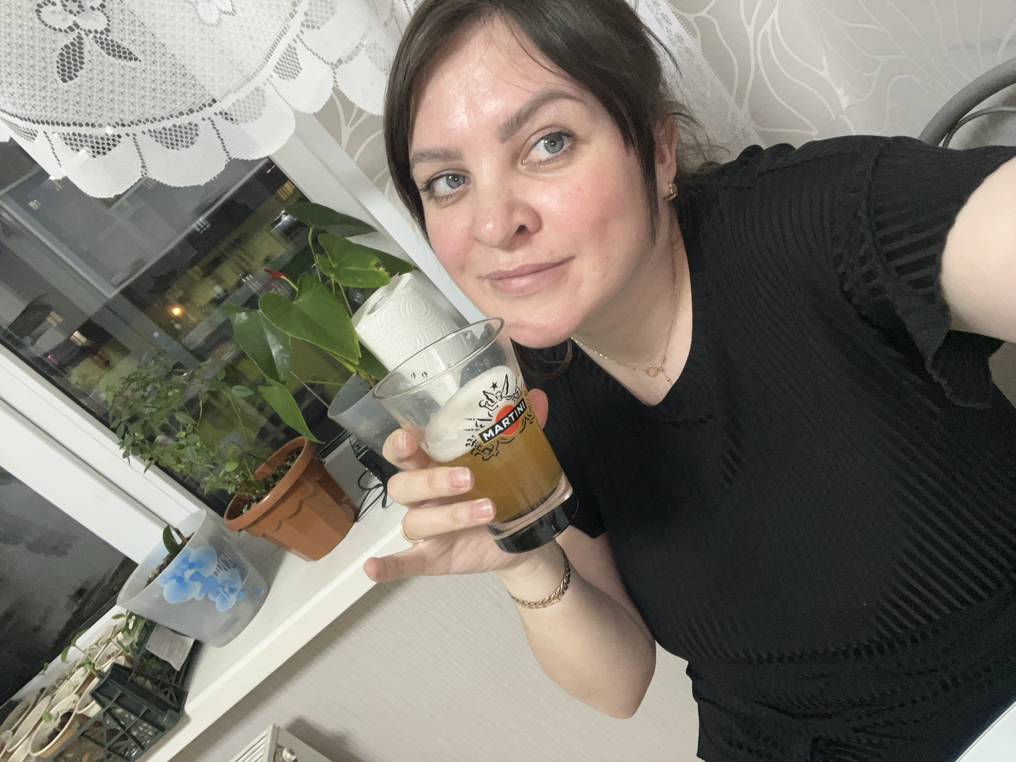 This indoor selfie captures a woman in Perm, Russia, holding a glass of a light amber beverage. The woman, appearing to be in her 30s or 40s with dark hair and blue eyes, looks directly at the camera with a faint, relaxed smile. She wears a black, vertically ribbed top or dress, a delicate gold chain necklace, and a gold-colored bracelet on her right wrist. Her nails have a natural, light polish.
The clear glass she holds features a prominent "MARTINI" logo, suggesting it might be a branded glass, although it contains what appears to be beer with a foamy head. Behind her, a window reveals a dark exterior with blurred street lights, indicating it is nighttime or late evening. The window sill is adorned with several potted plants, including one with large green leaves and a red bloom, and another in a pot with a blue floral design. A roll of paper towels is also visible on the sill. A white lace curtain hangs above the window, adding a domestic touch to the setting. The background wall features a subtle, patterned wallpaper. The overall scene suggests a casual and relaxed atmosphere.