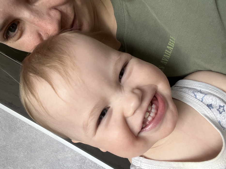 The image displays an indoor scene featuring a young child and the partial presence of an adult. A fair-skinned infant or toddler, with light-colored hair and blue eyes, is prominently positioned in the left and central foreground. The child exhibits a wide, open-mouthed smile, with squinted eyes and visible upper and lower teeth. The child is dressed in a white tank top patterned with light blue stars and other abstract shapes. To the right, only the adult's right facial profile, including a dark eye, and the upper torso are visible. The adult wears an olive-green t-shirt with light green text reading "IS AVAILABLE" on the chest. The child's head is positioned against the adult's upper body. The background consists of a light grey, possibly textured, wall with a vertical white trim or frame on the far left. No visual information within the image confirms a specific location in Chaikovsky, Russia.