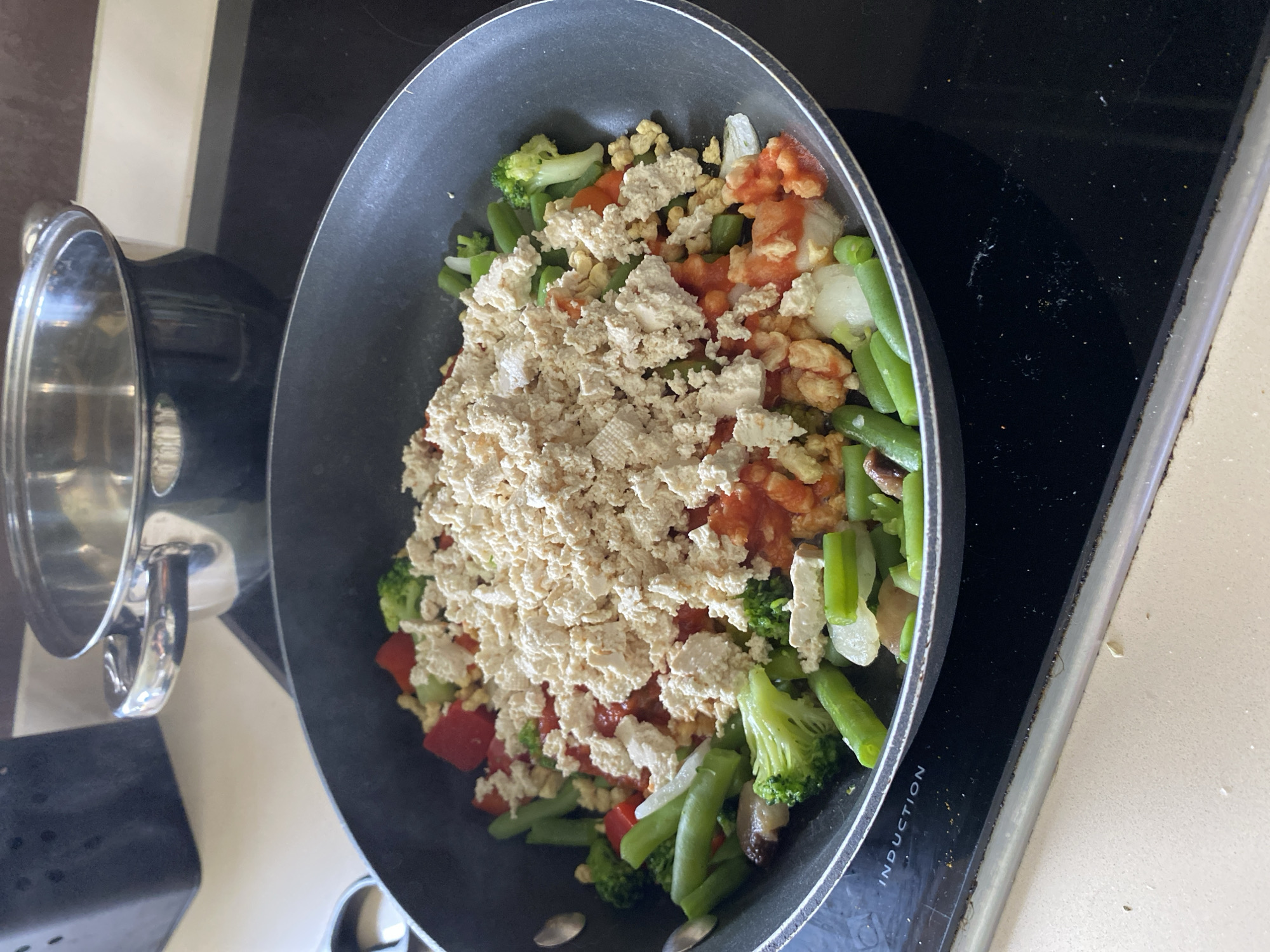 The image depicts a black frying pan on a black induction stovetop, likely in a kitchen. The pan contains a mixture of vegetables and what appears to be tofu. The vegetables include broccoli, green beans, red peppers, mushrooms, and onions. Tomato sauce is also visible. The tofu is crumbled and appears to be the main protein source in the dish. To the left of the frying pan is a stainless-steel pot with its lid off. The stovetop is on a light-colored countertop. The scene appears to be in a home kitchen. There is no indication of the specific city of Villaviciosa, Spain.