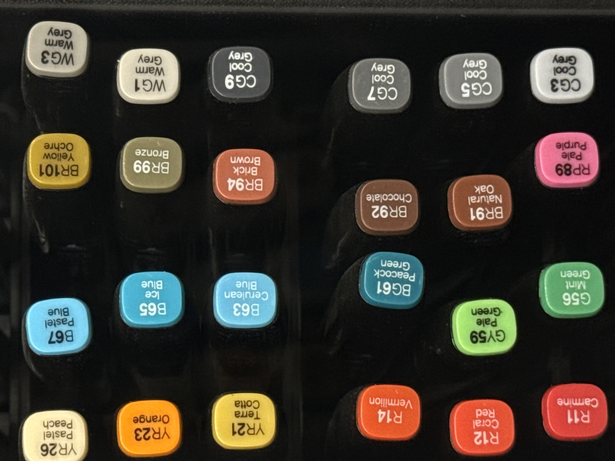 The image displays a close-up, top-down view of a set of art markers arranged in rows against a dark background. The markers have rounded caps with color swatches and labels indicating their color code and name.
In the first row, from left to right, are markers labeled "YR26 Pastel Peach", "B67 Pastel Blue", "BR101 Yellow Ochre", and "WG3 Warm Grey".
The second row features markers labeled "YR23 Orange", "B65 Ice Blue", "BR99 Bronze", and "WG1 Warm Grey".
The third row shows markers labeled "YR21 Terra Cotta", "B63 Cerulean Blue", "BR94 Brick Brown", and "CG9 Cool Grey".
The fourth row contains markers labeled "R14 Vermilion", "BG61 Peacock Green", "BR92 Chocolate", and "CG7 Cool Grey".
The fifth row displays markers labeled "R12 Coral Red", "GY59 Pale Green", "BR91 Natural Oak", and "CG5 Cool Grey".
The sixth row includes markers labeled "R11 Carmine", "G56 Mint Green", "RP89 Pale Purple", and "CG3 Cool Grey".
The image does not provide any information or visual cues that would allow for the determination of its location in the city of Moscow, Russia, or any other geographical context. The scene is a still life composition of art supplies. There are no people, actions, or interactions visible.