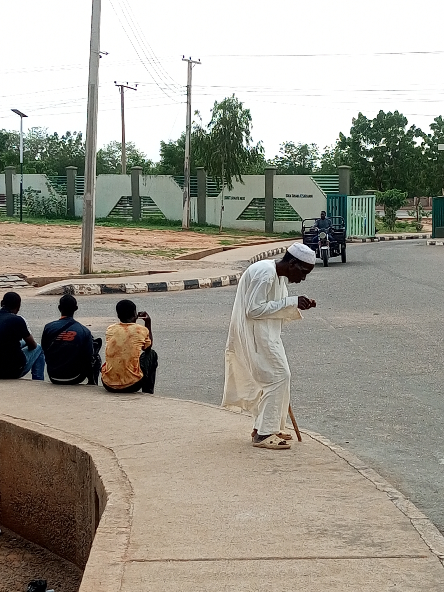 The image depicts an outdoor scene in Gombe, Nigeria. In the foreground, a man wearing a white traditional robe and cap walks with a cane, looking down at something in his hand. He is on a concrete sidewalk that curves along the edge of a paved road. Further down the sidewalk, three young men are sitting, facing away from the camera, with their backs to the road. One is wearing a dark blue t-shirt and jeans, another a dark blue shirt with an orange logo, and the third a yellow, patterned shirt.
In the background, there is a low concrete wall topped with a dark railing, separating the sidewalk area from a sandy, dirt area with some sparse vegetation. Behind this wall, a building with a pale green facade and horizontal dark green slatted accents is visible. Trees with dense green foliage line the top of this building and extend across the upper part of the image. Further in the background, a three-wheeled vehicle with a driver is moving along the road. The sky is overcast, suggesting a cloudy day.
Visible text on the wall reads "DON'T SMOKE HERE" on the left, and "BORNA SHARAMA STORE" on the right. The overall impression is a casual, everyday scene in a Nigerian town. The time of day appears to be daytime, given the light, but the overcast conditions prevent precise determination.