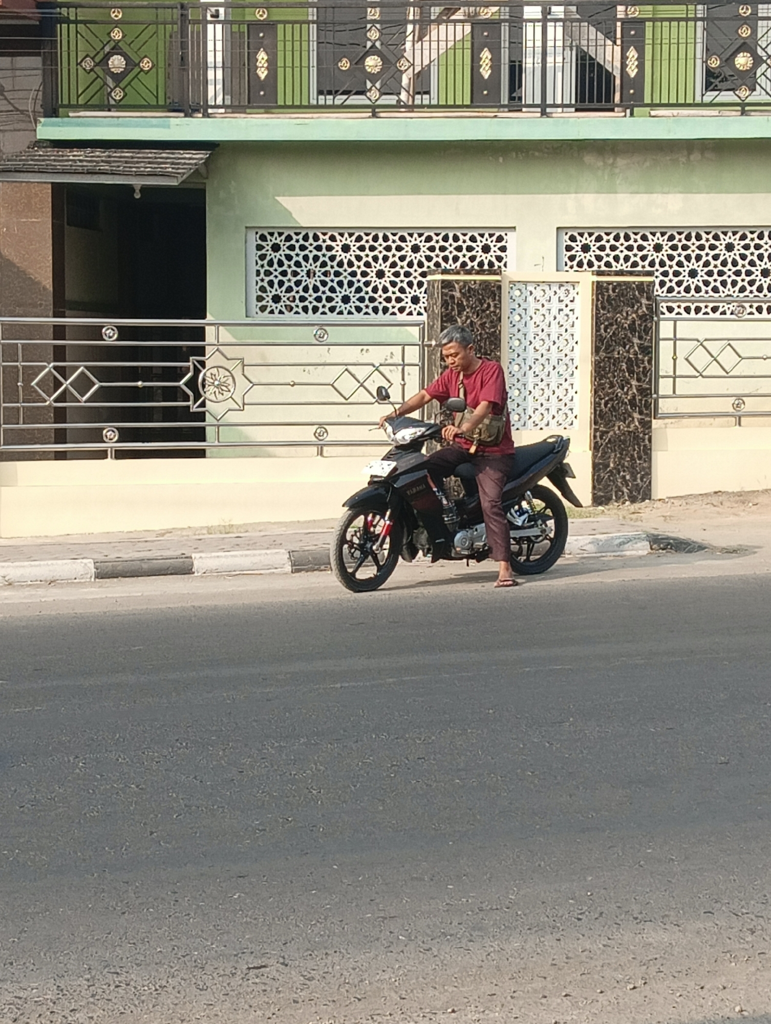 A man is sitting on a black Yamaha motorcycle in front of a pale green building. He is wearing a maroon t-shirt, dark maroon pants, and flip-flops. A tan bag is slung across his chest. The building has decorative metalwork and patterned windows. A street with a curb and sidewalk is in the foreground, and the motorcycle is on the street. The location appears to be Leuweunggajah, Indonesia, based on the context provided, though this cannot be definitively confirmed from the image itself. The environment suggests an urban or suburban setting during daylight hours.
