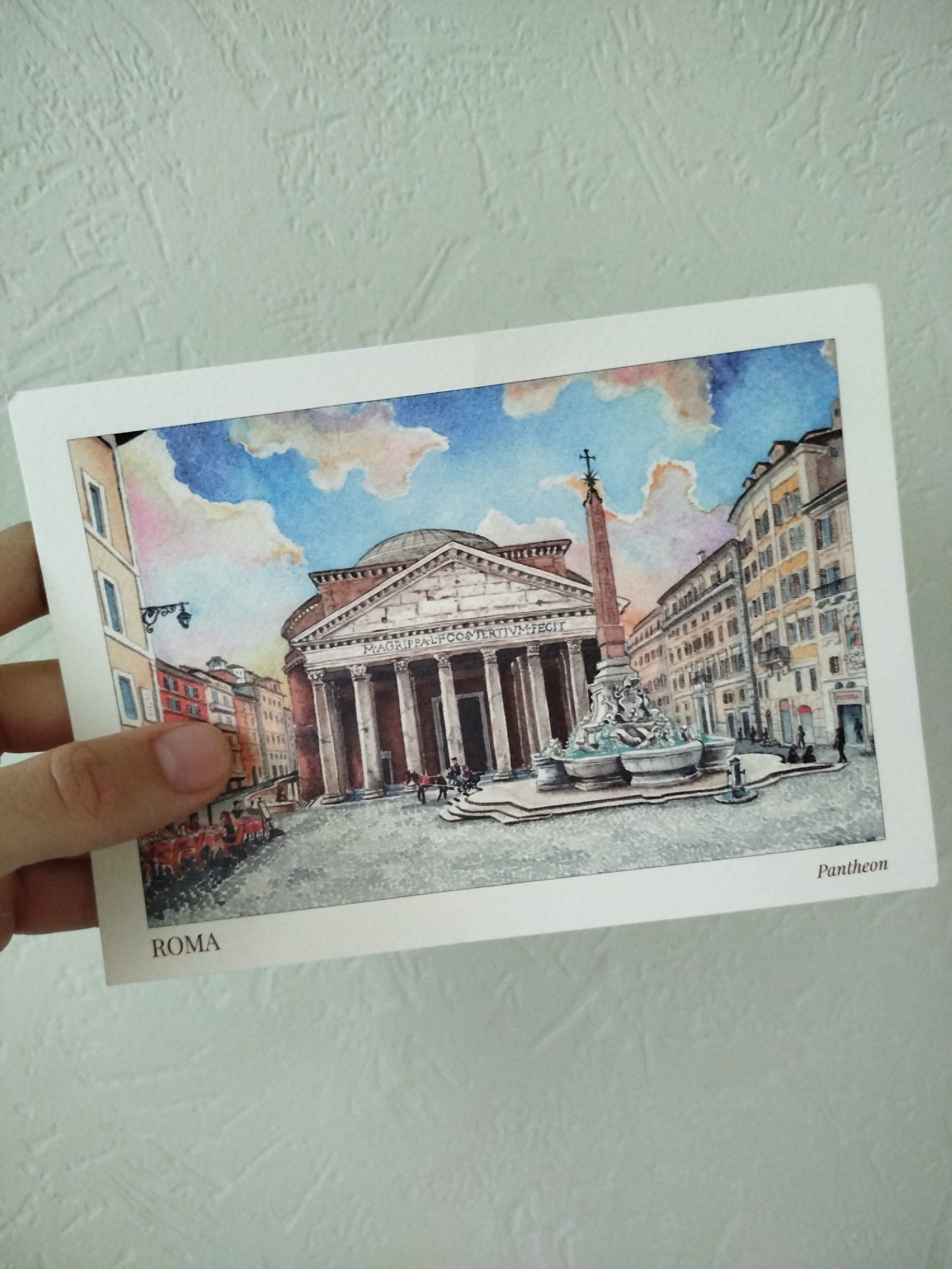 A hand holds a postcard depicting the Pantheon in Rome, Italy. The watercolor painting on the postcard features the Pantheon's facade with its columns and dome, an obelisk atop a fountain in the piazza, surrounding buildings, and a blue sky with clouds. The word "ROMA" is printed at the bottom left of the postcard, and "Pantheon" is printed at the bottom right. The background is a textured white wall.