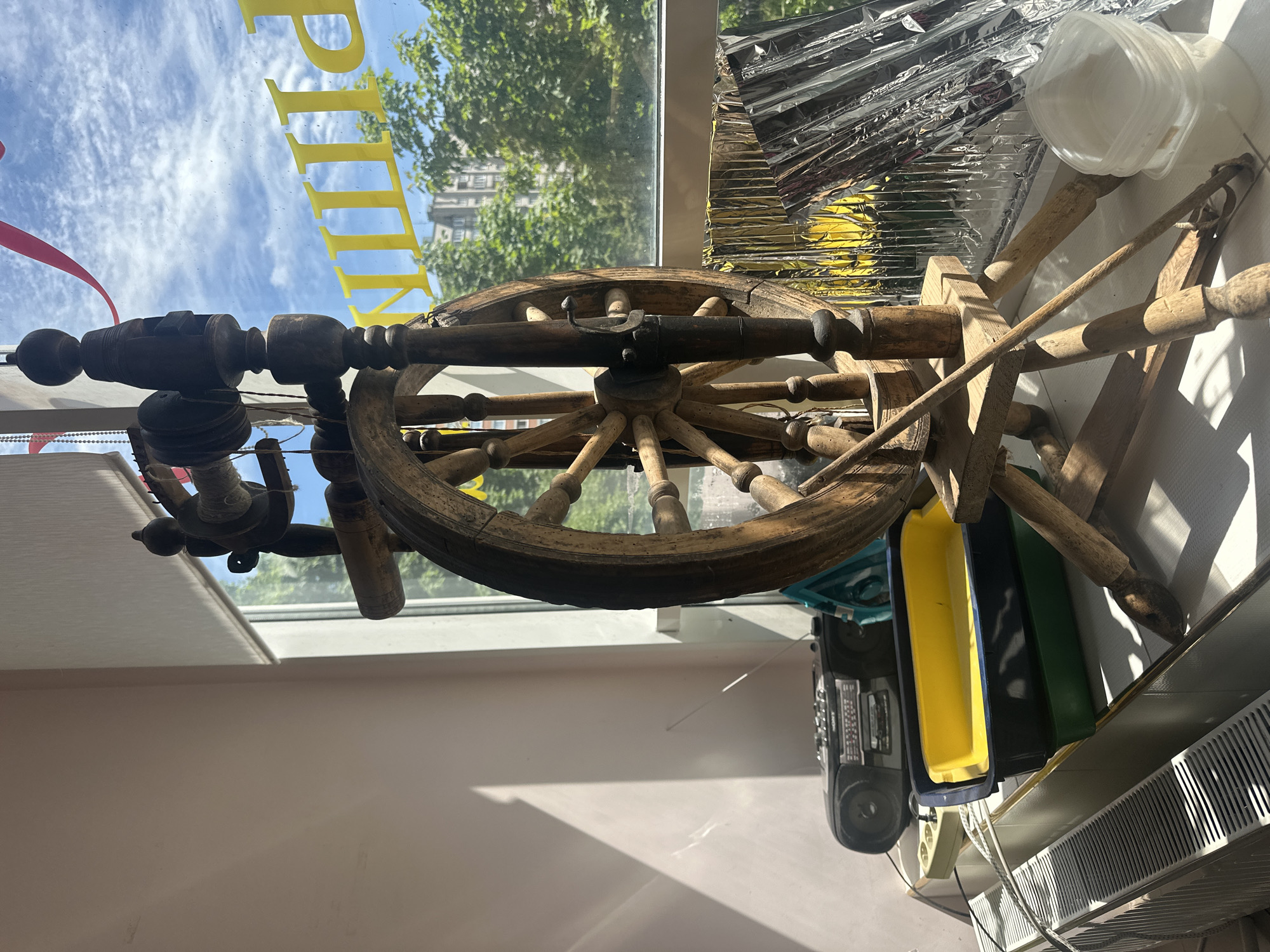 The image displays an indoor scene looking out through a window during daytime. A large, antique-style wooden spinning wheel dominates the foreground, positioned centrally in front of the window. The spinning wheel features an intricate spoked wheel, various turned wooden components, and a bobbin/flyer mechanism on its left side. A red ribbon hangs to the left of the spinning wheel.
Through the window, a clear blue sky with scattered white clouds is visible, along with the canopy of green trees and the facade of a distant multi-story building, suggesting an urban or suburban setting in Stavropol, Russia. Yellow block letters are affixed to the window, viewed from inside, clearly showing the partial word "PITTIN" as if reversed from an outdoor sign.
To the right of the spinning wheel, near the window frame, several clear plastic containers are stacked. Below the window sill and to the right, various objects are present: crumpled silver foil or insulation material, a vintage-style boombox/radio, and three colorful plastic trays (yellow, blue, and green). A white power strip and a white radiator with horizontal fins are also visible along the bottom right.
Sunlight streams through the window, casting distinct shadows on the interior surfaces, confirming it is daytime. No people or activities are depicted; the scene is static.