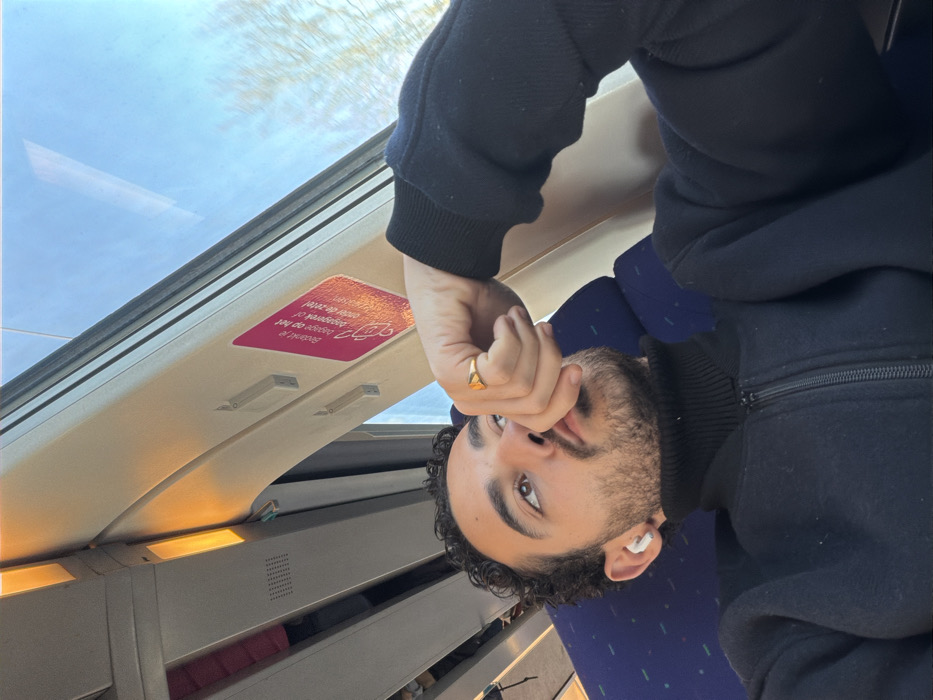 The image displays a male individual seated inside a train carriage, identified by context as being in the city of Chauny, France. The subject has short, dark, curly hair, a beard, and a mustache. He wears a black high-neck zippered garment. A white earbud is inserted into his left ear. His right hand is raised, with fingers positioned near his mouth, and a gold-colored ring is visible on the ring finger of this hand. He is looking upwards and to his right. The train interior includes blue patterned seats and white overhead panels with some illuminated orange-yellow light fixtures. A pink rectangular sticker with Dutch text, "Bedankje op het dagmenu of onze de zetel!", is affixed to the overhead panel near the window. Outside the window on the right, a clear blue sky and bare tree branches are visible, indicating daytime and a season without foliage. The background elements appear slightly blurred, consistent with the movement of the train.