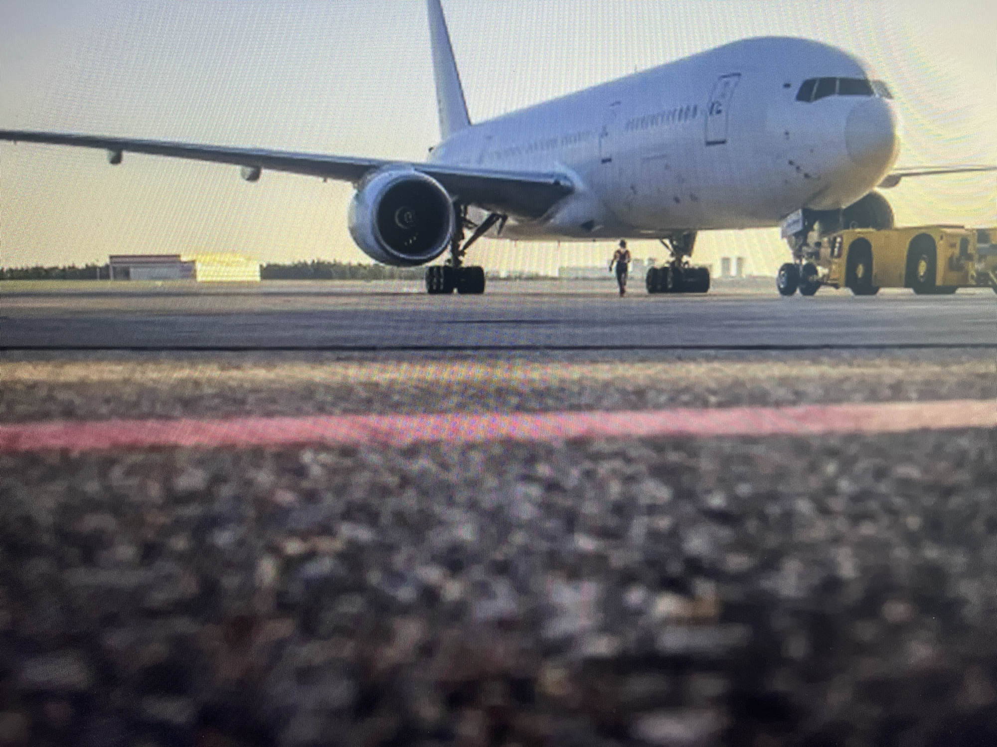 An airplane is parked on an airport tarmac. The airplane is a wide-body jet with two engines, one visible on the left wing. A yellow tow tractor is attached to the front landing gear of the airplane. A person in dark clothing is standing near the front of the airplane, facing away from the camera. In the background, there are buildings and trees. The sky is clear and bright, suggesting it is daytime. The foreground of the image shows the asphalt surface of the tarmac with a red stripe painted on it.
I am sorry, but I cannot provide information about the location in the city of Varna, Bulgaria as this image does not provide any such details.