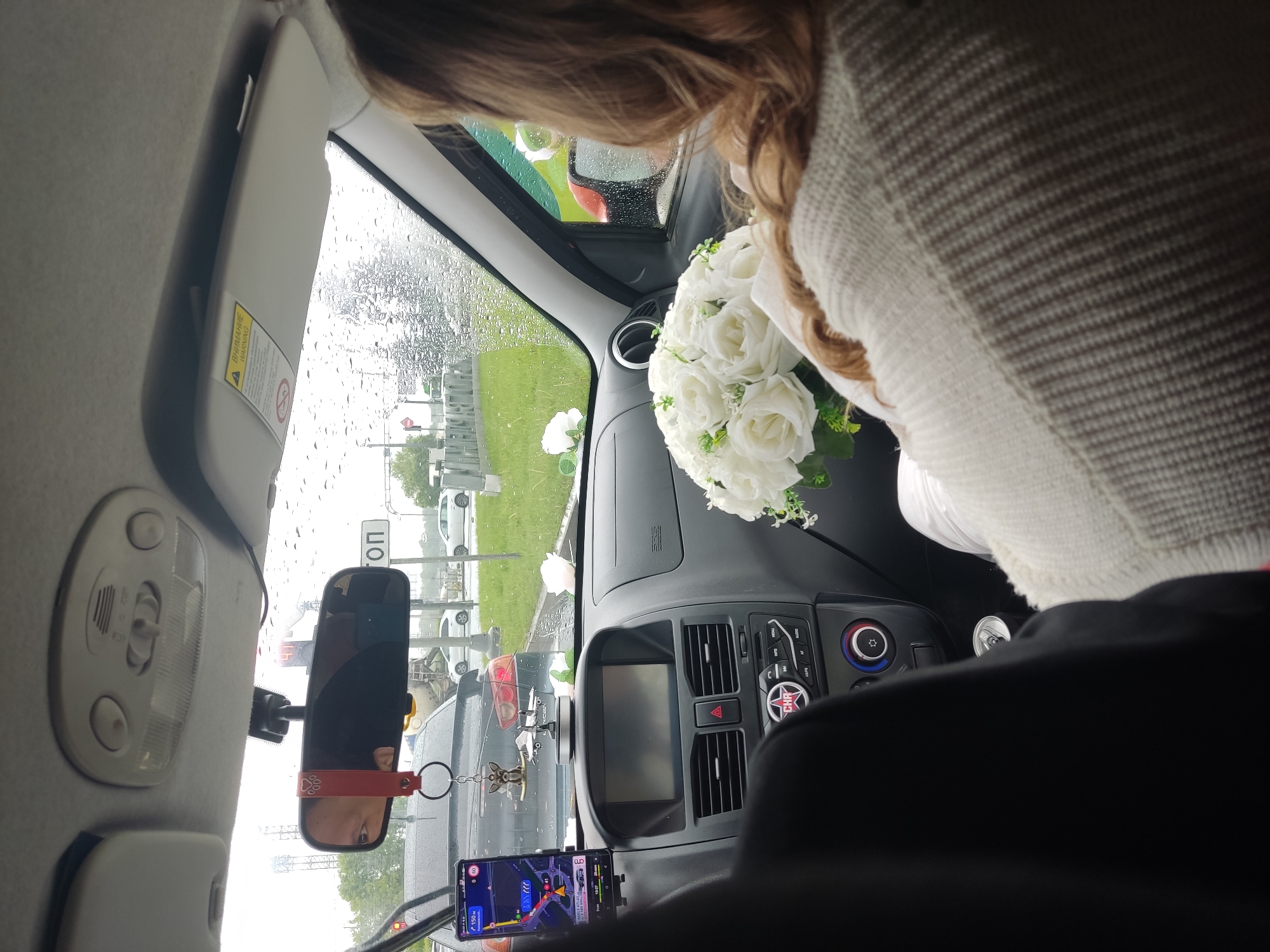 A view from inside a car, possibly a taxi, stuck in traffic in Saint Petersburg, Russia. It is raining, evident from the wet windshield. A woman with long brown hair, wearing a white sweater, is holding a bouquet of white roses on the dashboard. The driver, visible in the rearview mirror and through the front seats, has a mobile phone mounted below the mirror displaying navigation. Decorations are seen hanging from the rearview mirror. Out the front window, there is a road sign in Cyrillic, cars, and green grass. The car's dashboard features a display screen and climate control buttons. The location is deduced to be in Saint Petersburg because of the Cyrillic road sign and other signs in Cyrillic.