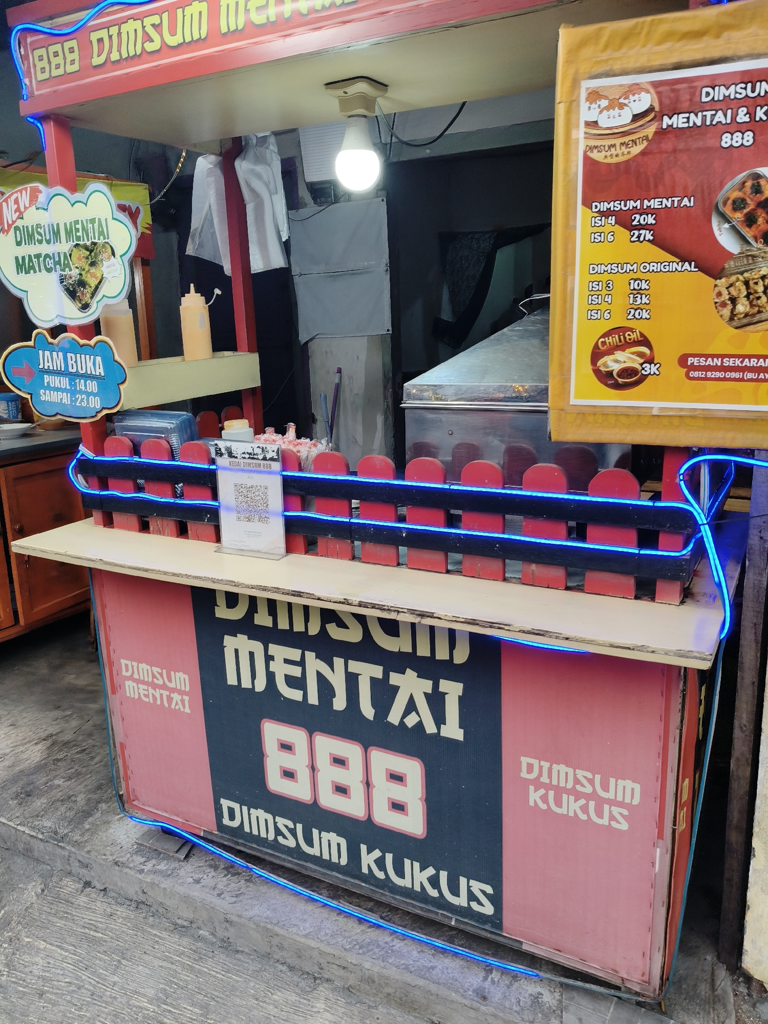The image captures a dim sum food stall with prominent branding. The stall is located at an outdoor market or sidewalk in South Tangerang, Indonesia. The stall features a red and yellow canopy with "888 DIMSUM MENTAI" written in large, stylized text. A sign on the left advertises "NEW DIMSUM MENTAI MATCHA" and the stall's operating hours are listed as "JAM BUKA PUKUL: 14.00 SAMPAI: 23.00". A yellow sign on the right displays pricing for "DIMSUM MENTAI" and "DIMSUM ORIGINAL", as well as "Chili Oil" for 3K. The stall has a decorative red and black fence with blue neon lights. In the background, there is kitchen equipment and what appears to be stacks of boxes. The ground surface is paved with concrete. There are no people visible in the image, and no visible actions or interactions are taking place.