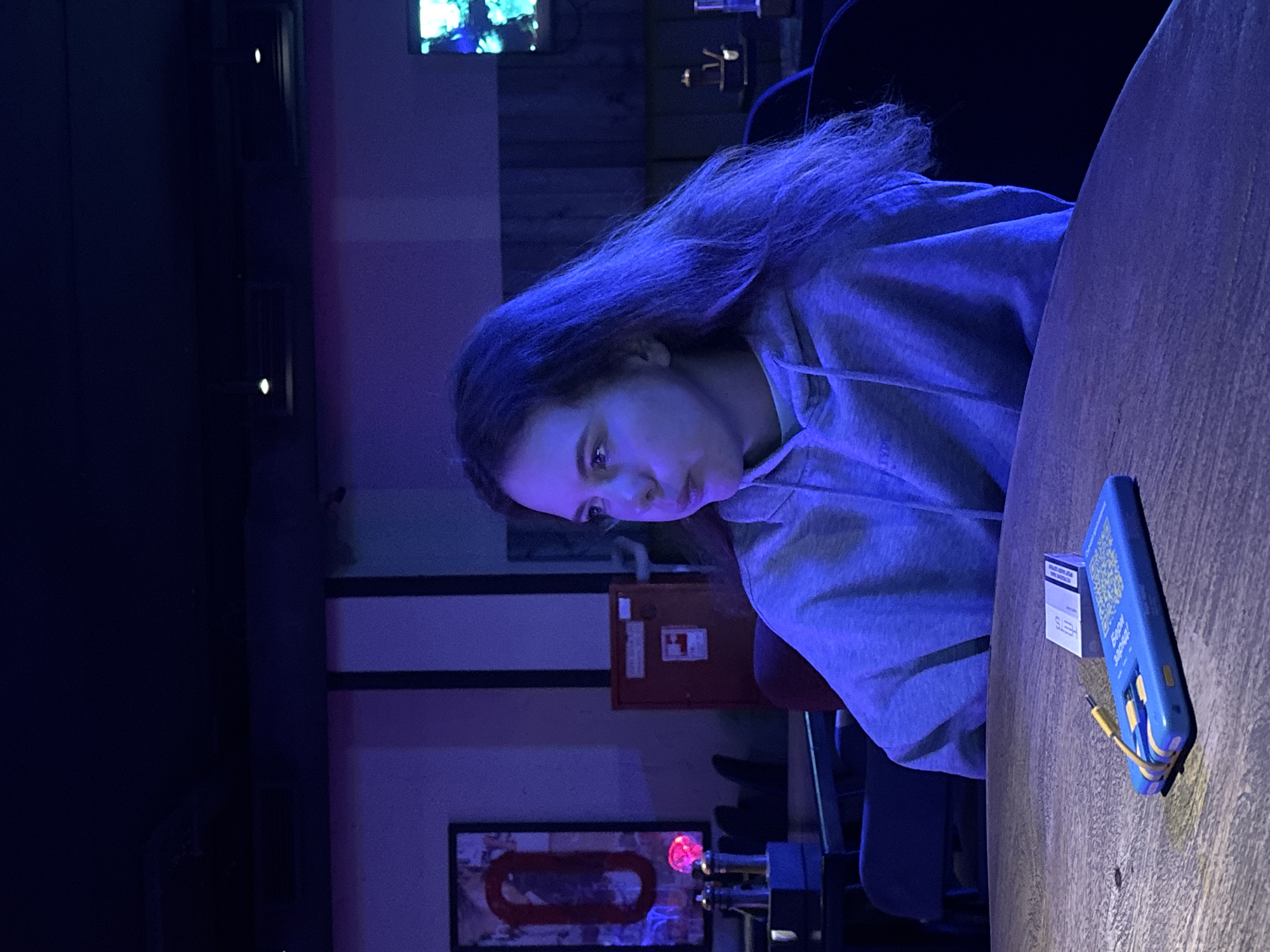 A person with long hair, wearing a light-colored hoodie, sits at a wooden table. A blue phone case with a yellow cord and a pack of cigarettes are on the table. The scene is lit with blue light. A painting is visible on the wall and a fire extinguisher box. The location is Krasnoyarsk, Russia.