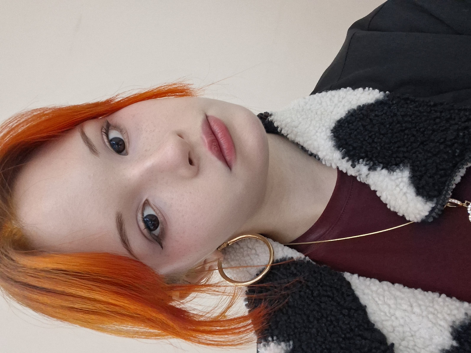 A close-up, angled portrait shows a person with dyed, orange hair. The individual is wearing a maroon-colored shirt and a jacket with a black and white, shaggy fur collar. They are also wearing a gold hoop earring and a delicate gold necklace with a small, possibly bejeweled, pendant. The person has fair skin, visible freckles, and wears subtle eyeliner. The background is a neutral, light beige color. There is no information about the location being in Chaikovsky, Russia, in the image.
