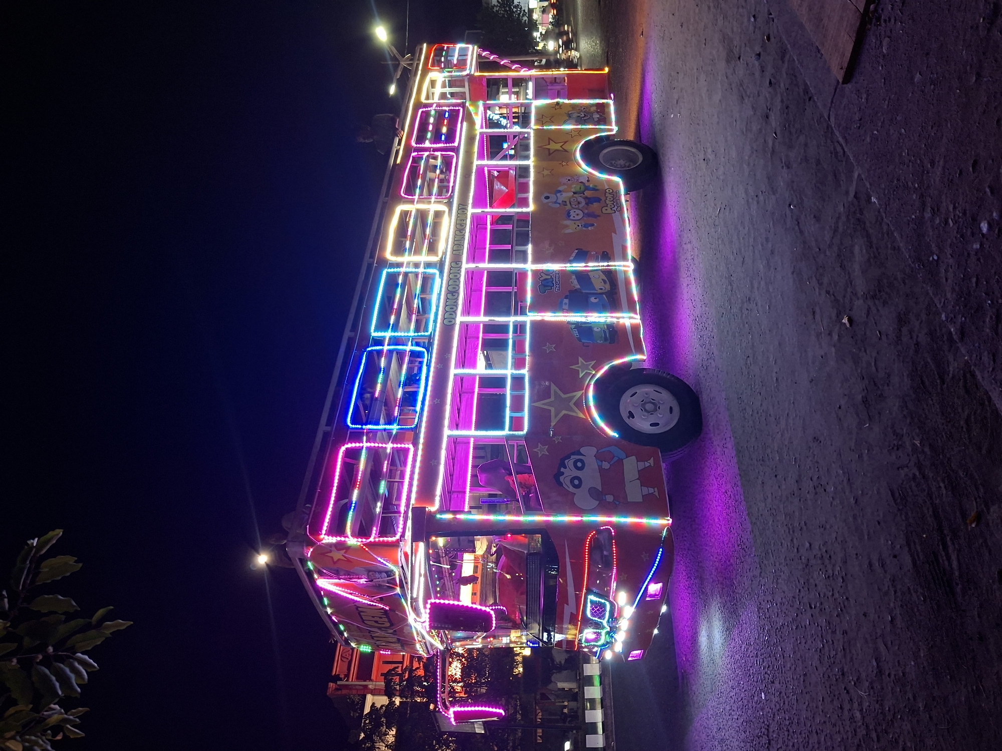 This image captures a vibrant, customized double-decker bus illuminated by colorful LED lights at night in Lhokseumawe, Indonesia. The bus, likely an "Odong-odong" or mobile amusement ride, is stationary or moving slowly on a dark road.
The bus itself is a prominent red-orange color, heavily adorned with bright LED strip lights in magenta, blue, yellow, and green, outlining its windows, body panels, and wheels. These lights cast colorful reflections on the asphalt or dirt road below, creating a festive atmosphere. Cartoon characters popular with children, such as Shin-chan, Pororo the Little Penguin, and Tayo the Little Bus, are prominently painted on its sides, alongside star motifs.
Visible text on the bus includes "ODONG ODONG ADIK GENOY" on the upper deck and "TAYO" and "Pororo Coo coo" on the lower deck, further indicating its function as an entertainment vehicle. The dark sky in the background confirms it is nighttime, with distant streetlights hinting at an urban or semi-urban setting. Silhouettes of people, possibly passengers or operators, can be seen on the bus's upper deck, with one figure holding a light source. A plant with broad leaves is visible in the lower left foreground. The overall scene exudes a playful and lively energy.