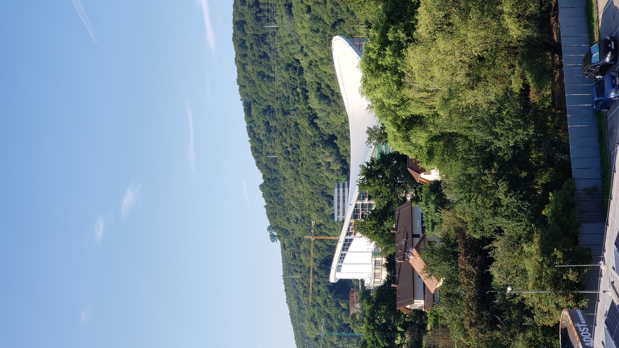 The image depicts a modern building under construction, characterized by its large, curved, white roof. The structure is situated on the outskirts of Prešov, Slovakia, with a densely wooded hill serving as a backdrop. Several cranes are visible around the construction site, indicating ongoing work. In the foreground, several residential houses with red-tiled roofs are nestled among lush green trees. A street light is also visible in the lower left of the image. Further to the left, a circular sign with the partial text "ADOST RESTAURAC" is present, suggesting a commercial establishment nearby. Two cars, a blue sedan and a black hatchback, are parked in a lot in the bottom right corner of the image. The sky is a clear, pale blue with a few wispy clouds. The overall scene suggests a suburban or semi-rural setting on a sunny day.