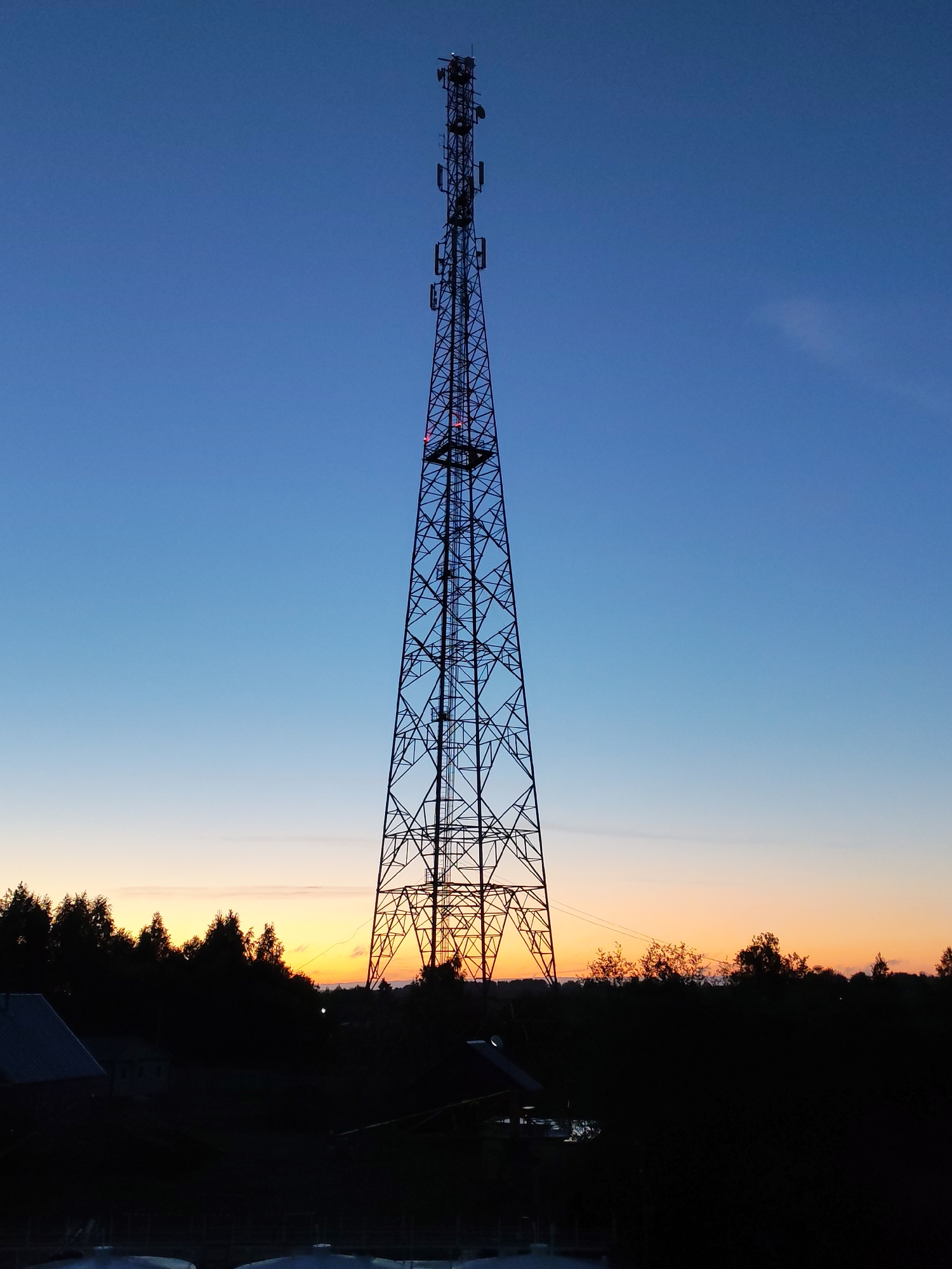 A low-angle shot depicts a tall metal lattice telecommunications tower silhouetted against a gradient sunset sky. The sky transitions from deep blue at the top to shades of orange and yellow near the horizon. The tower extends high into the frame, with antennas and other equipment visible near the top. The base of the tower is situated behind a dark treeline and a building.
The image was taken in Annensky Most, Russia.