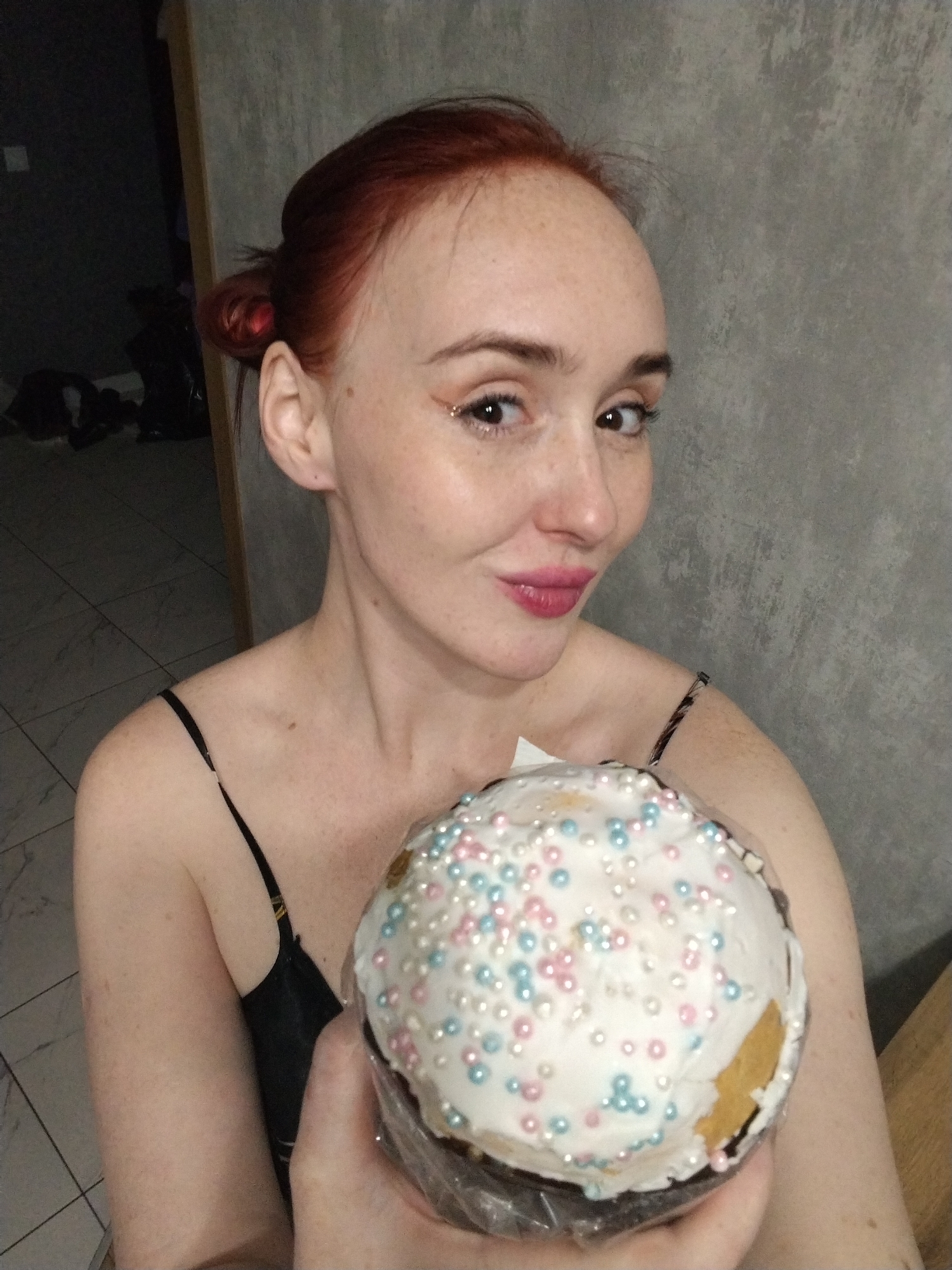 A fair-skinned, red-haired woman poses indoors, holding a decorated cake. The woman has freckles, light eye makeup, and pink lipstick. Her hair is pulled back into a bun. She wears a black tank top with thin straps. The cake is frosted with white icing and decorated with pink, white, and blue sprinkles. The location is unknown and there is no information related to Izhevsk, Russia, in the image. The background consists of a gray wall and a dimly lit hallway with objects on the floor. The woman is holding the cake in front of her.