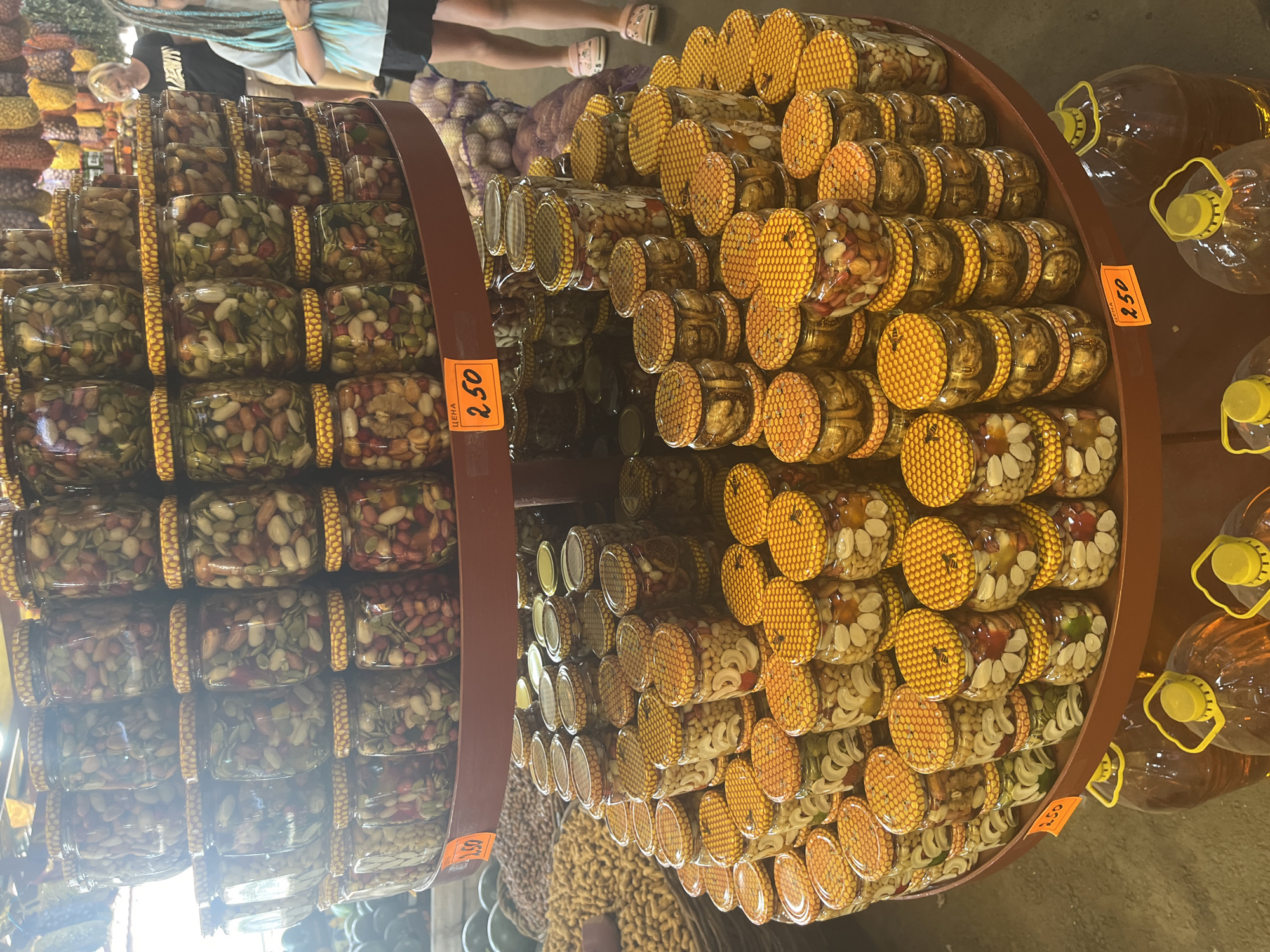 The image depicts a market stall in Plastunovskaya, Russia, displaying a large assortment of preserved goods in glass jars, primarily nuts and seeds. The jars are arranged in stacks on a tiered circular display shelf. The lids of many jars are decorated with a honeycomb pattern, and some feature small bee illustrations. In the foreground, large plastic bottles filled with a dark amber liquid, likely oil, are visible. In the background, two young females, one with blue braided hair, are partially visible, along with other market stalls and produce. An orange price tag indicating "2.50" is attached to the display shelf. The overall scene suggests a vibrant outdoor or indoor market environment.