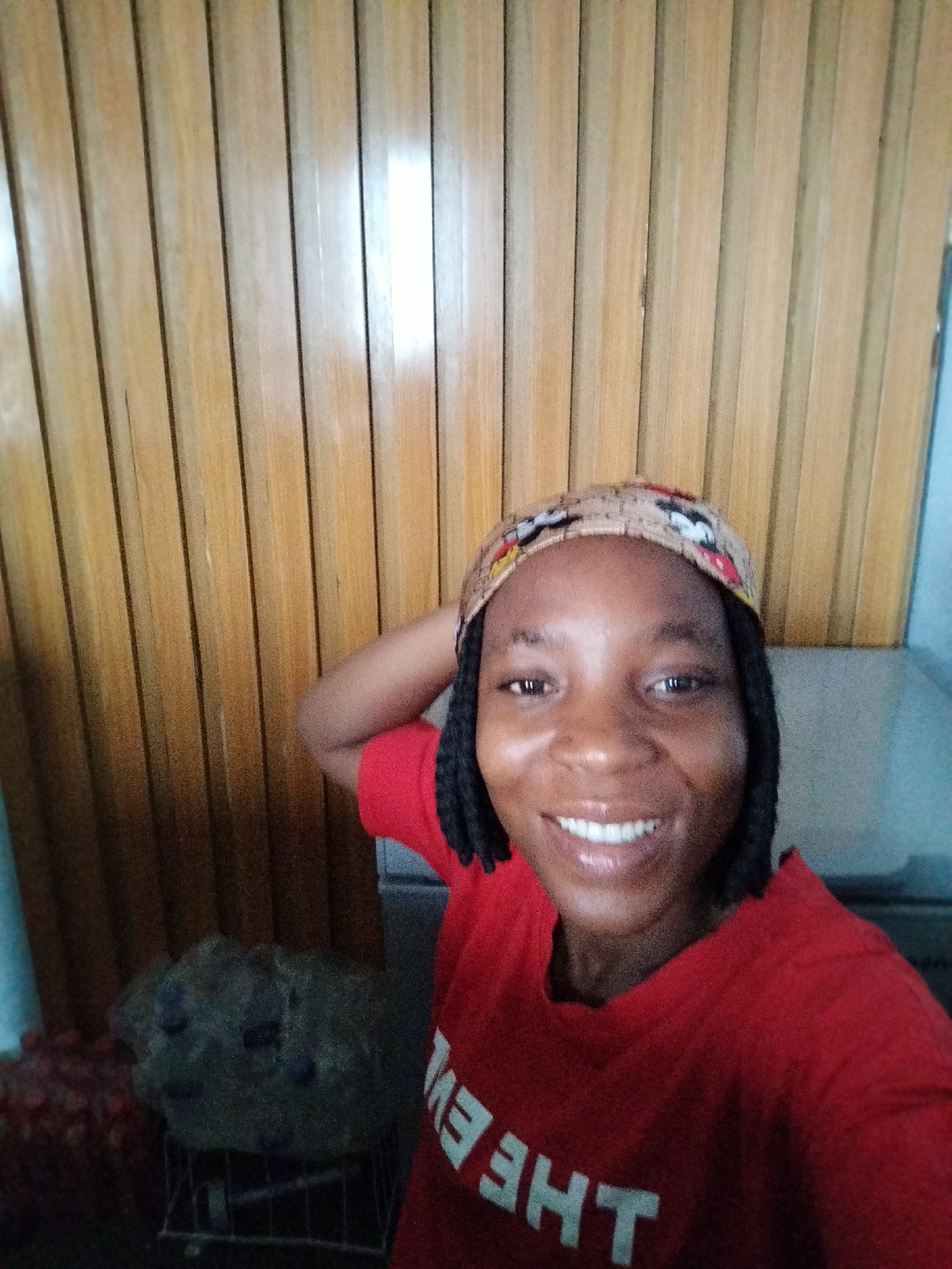 A young woman wearing a red t-shirt and a patterned headwrap is posing for a selfie. She is smiling and has her arm raised. Behind her is a wall of vertical wooden planks. To the left of the woman, partially visible, is a shopping cart with some items in it. The scene appears to be indoors. The lighting is somewhat dim, but the woman's face is well-lit.