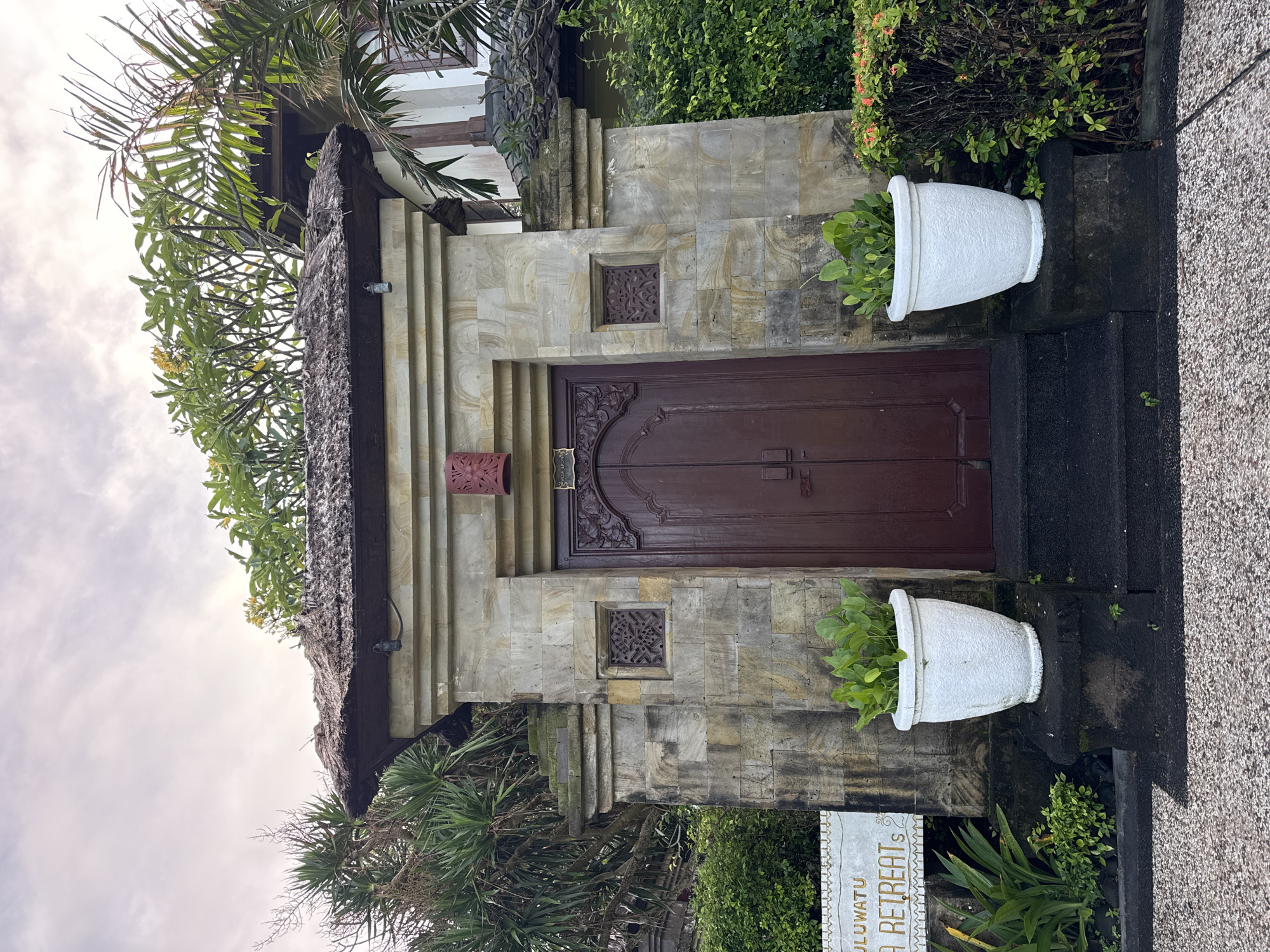 The image displays a traditional Balinese-style entrance, likely for a private villa or retreat, set within a lush tropical environment. A large, dark brown double wooden door, intricately carved with floral and swirling motifs, serves as the central focal point. It is framed by walls constructed from variegated light grey and beige stone tiles, exhibiting natural patterns and textures.
Above the door, two smaller, square, decorative carved panels are integrated into the stone facade, mirroring the ornate style of the main door. A smaller, red-orange carved element is affixed to the stone wall to the left of the door. A thatched roof, characteristic of Balinese architecture, extends over the top of the entrance.
Two large, white, urn-shaped planters, each holding vibrant green leafy plants, flank the entrance, sitting atop dark stone pedestals. The ground in the foreground consists of dark textured steps and a lighter, speckled pathway. Surrounding the architectural structure, dense tropical foliage, including various green bushes, palm fronds, and some plants with small yellow flowers, thrives under an overcast sky with soft white clouds, indicating daytime.
On the lower right side, a white sign board with golden decorative elements is partially visible, displaying the text "ULUWA... A RETREATS". This suggests the location is a retreat, possibly named 'Uluwatu Retreats', aligning with the provided Kuta Selatan, Indonesia context. The scene is devoid of people, emphasizing the serene and private nature of the entrance.
