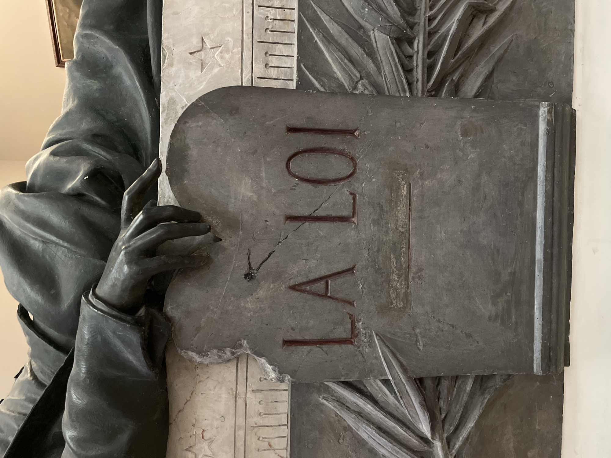 The image displays a close-up, low-angle view of a detail from the Monument to Victor Noir, located in Père Lachaise Cemetery, Paris, France. The primary subject is the dark-patinated bronze right arm and hand of a reclining male effigy, resting upon a weathered, dark gray stone tablet. The sculpted hand is extended with fingers slightly curled, pointing towards the upper right corner of the tablet. The tablet, irregular in shape with a fractured upper-left edge and visible cracks traversing its surface, bears the deeply carved inscription "LA LOI" (The Law) in prominent red capital letters. A recessed horizontal rectangular indentation is present below the text. The tablet is supported by a lighter, off-white marble or stone plinth, adorned with relief carvings including a distinct five-pointed star on the right and stylized, overlapping fronds or branches. A plain, light-colored background wall is visible behind the monument.