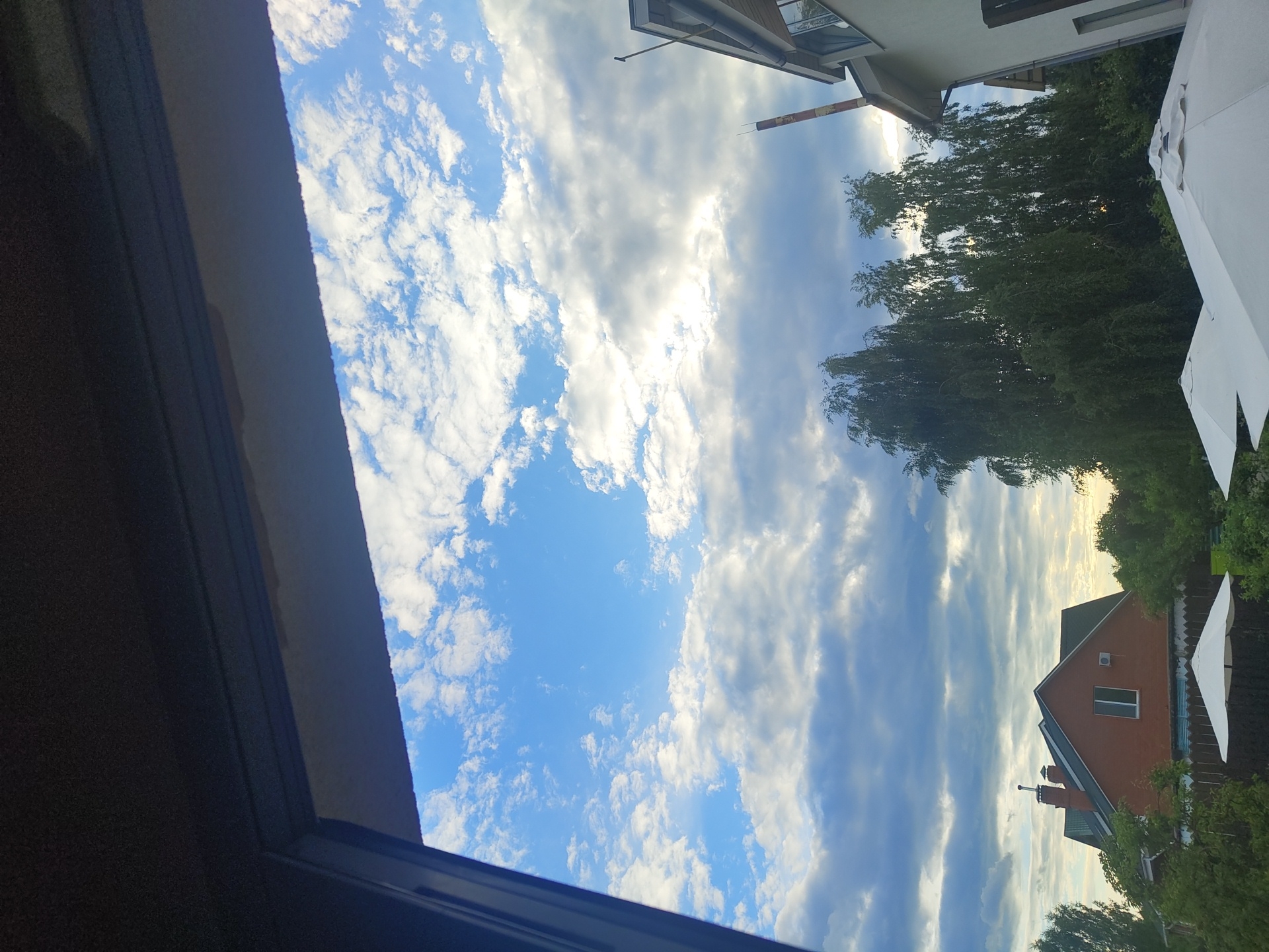 The image depicts an outdoor scene viewed through a window frame, looking out towards a residential area under a cloudy blue sky. In the foreground, partially obscuring the view, are the dark, textured surfaces of the window frame, likely made of wood or a similar material.
Beyond the window, the sky is predominantly blue with scattered white and gray clouds. The clouds vary in density, with some appearing wispy and others more substantial, suggesting a dynamic weather pattern. Sunlight is visible breaking through some of the cloud cover, creating brighter areas in the sky.
In the mid-ground, several residential buildings and trees are visible. To the left, a two-story house with a red brick exterior and a dark pitched roof is prominent. It features a chimney and a single visible window. Surrounding this house and extending across the scene are lush green trees, some of which are tall and leafy, possibly birch trees due to their slender appearance.
To the right of the frame, a portion of another building, possibly a two-story house with a light-colored facade and a dark roof, is visible. A tall, cylindrical structure, resembling a chimney or a pipe with a reddish-brown hue and white bands, extends upwards behind the trees.
In the foreground, at the bottom of the image, two white umbrella-like structures are visible, likely patio umbrellas, suggesting an outdoor seating or recreational area.
The scene appears to be set in a suburban or rural residential area, consistent with the location of Цибино, Russia. The overall impression is a pleasant, possibly late afternoon or early evening, view from indoors looking out at a tranquil outdoor environment. There are no people or discernible actions captured in the image.