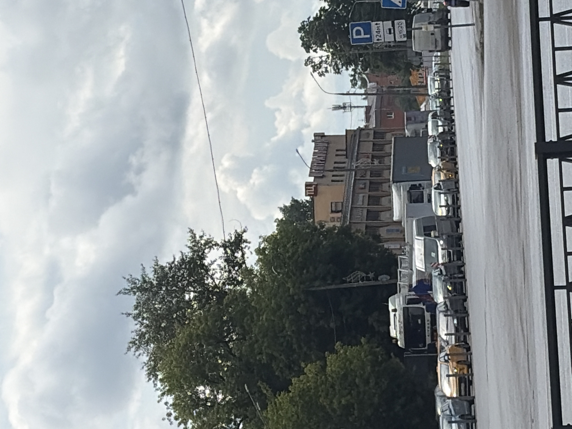 The image depicts a traffic scene on a city street in Podolsk, Russia, under a cloudy sky. In the foreground, a gray road surface is visible, with a black metal barrier in the lower portion. A line of cars and vans is stopped on the road, appearing to be in traffic. Among the vehicles are several cars, a yellow taxi, and larger trucks, including a white truck with "AITMAN" written on the front, and a gray container truck.
To the right of the vehicles, a blue and white parking sign is mounted on a pole. Above the vehicles, large green trees with dense foliage are prominent, with some branches extending towards the sky. Behind the trees and to the right, a multi-story building with a brick facade and numerous windows is visible. In the distance, a telecommunication tower with antennas can be seen. The overall environment suggests an urban setting with significant vehicle presence.