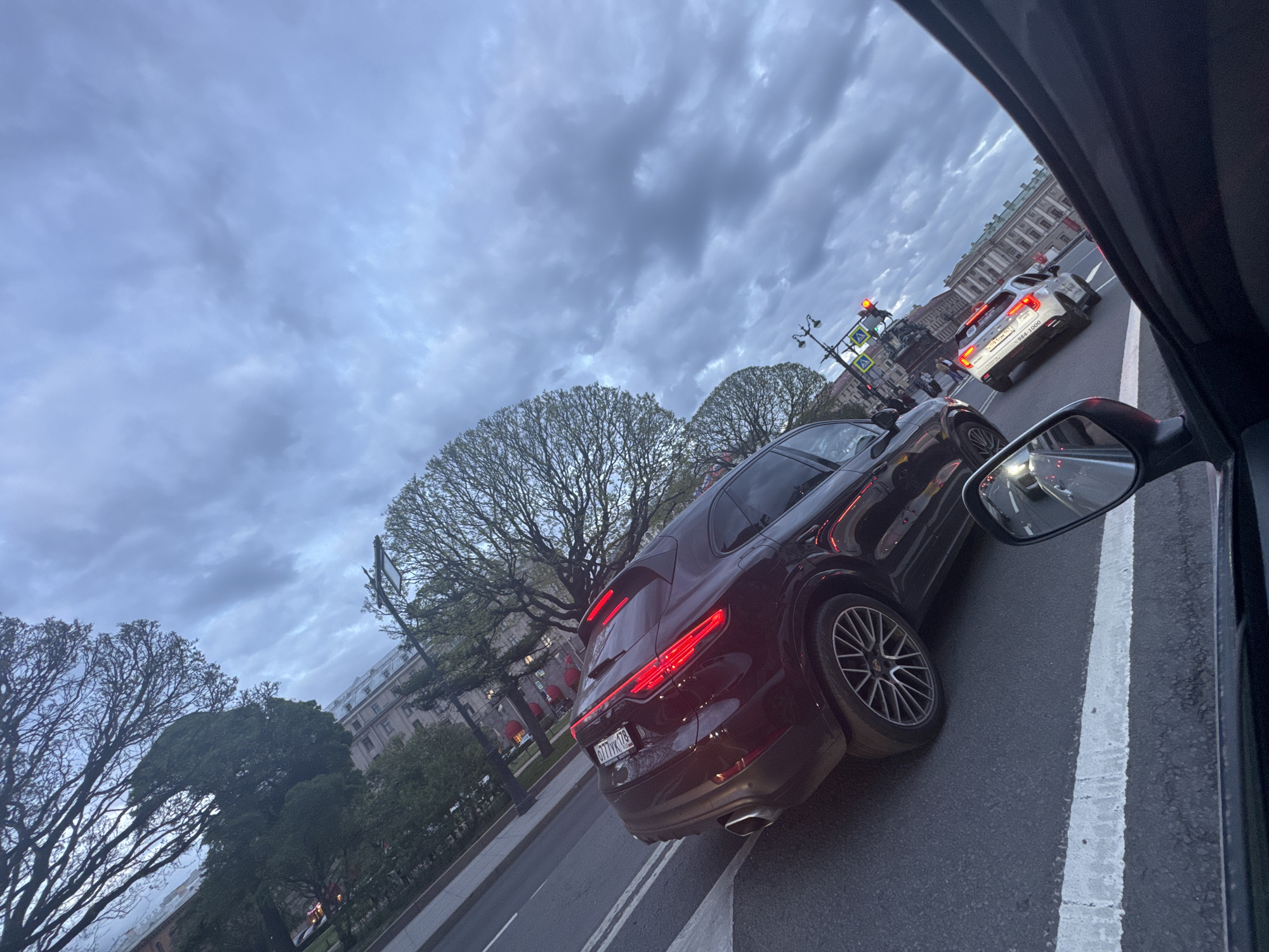 The image shows a street scene in Saint Petersburg, Russia. The photograph was taken from the interior of a car on a city street. A dark-colored Porsche Cayenne is adjacent to the photographer's vehicle. Behind the Cayenne is a white car. A street light with traffic lights are visible, and pedestrian crosswalks. Buildings line the opposite side of the street, and trees are interspersed between the buildings. The sky is overcast with dark clouds.