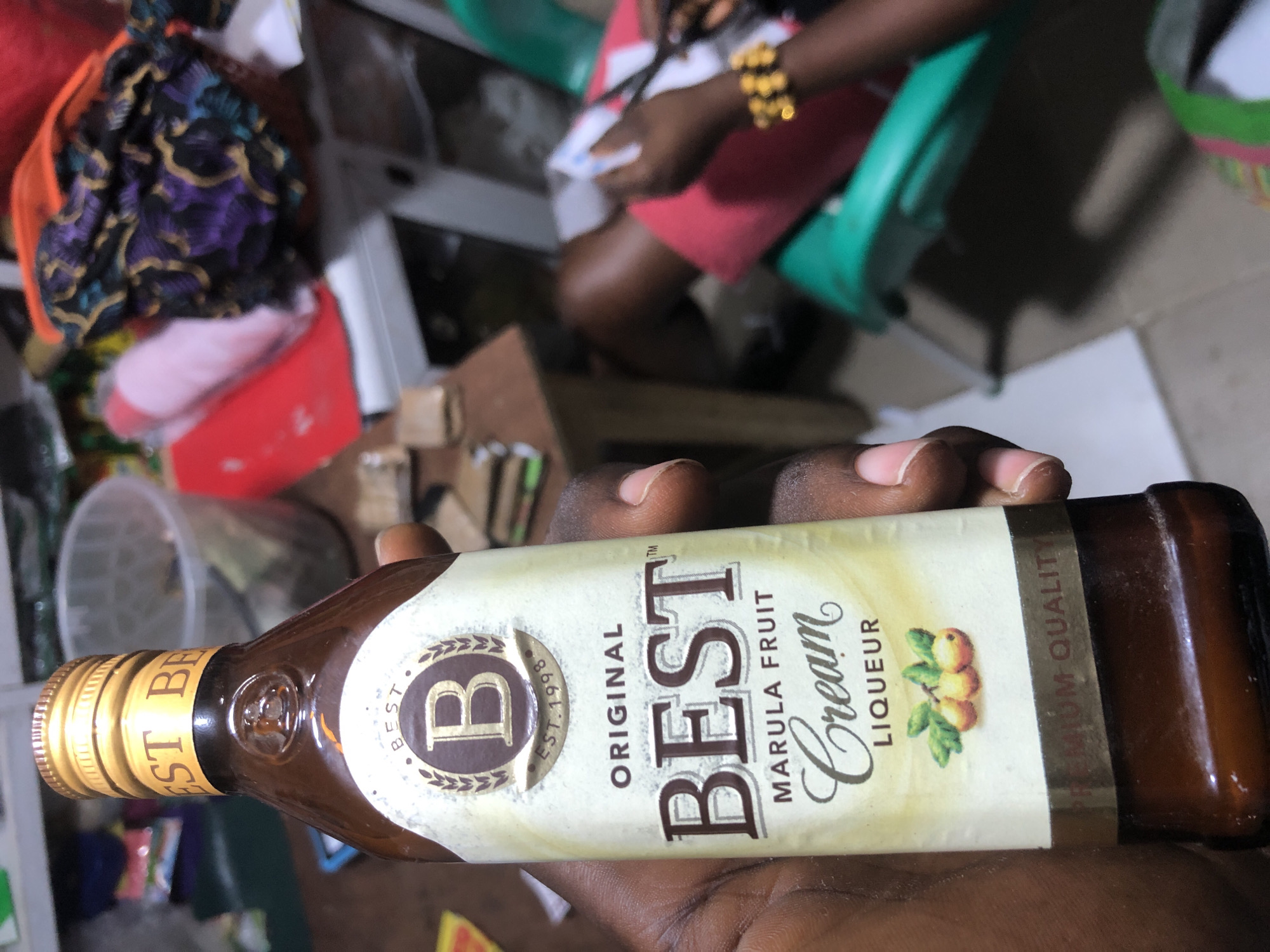 An individual is holding a bottle of "BEST Original Marula Fruit Cream Liqueur" in Ado, Nigeria. The bottle is brown glass with a gold label featuring a large "B" logo and the text "BEST MARULA FRUIT Cream LIQUEUR". The label also indicates "EST. 1998" and "PREMIUM QUALITY". In the background, there are other items on a table, including fabrics, a pink object, and a transparent container. Another person is visible in the background, wearing a gold bracelet and holding a pair of scissors, appearing to be cutting a piece of paper. The environment suggests an indoor setting, possibly a shop or a home.