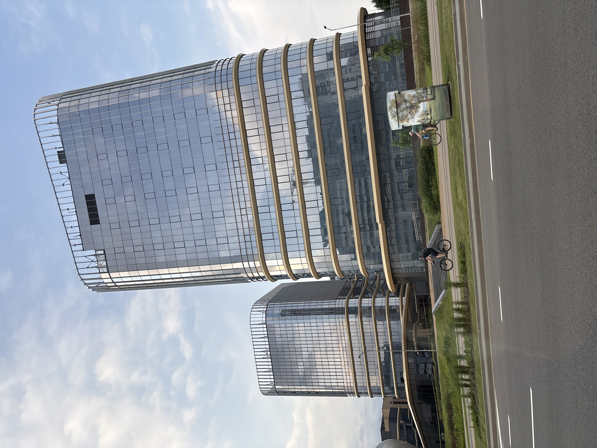 The image shows two modern, glass-facade office buildings with curved designs and golden horizontal accents. The taller building is on the right, and a slightly shorter one is on the left. Both buildings have a metal railing structure at the top. In the foreground, a paved road runs horizontally across the bottom of the frame, with white lane markings. A green grassy area with some bushes is situated between the road and the buildings. Two individuals are visible cycling on a path within this grassy area. One cyclist, wearing a dark t-shirt and shorts, is on the right side of the frame, riding a bicycle. Another cyclist, in a light-colored t-shirt and shorts, is further to the left. A rectangular outdoor advertisement displaying a landscape with trees is positioned near the right-hand cyclist. The sky is partly cloudy with a mix of blue sky and white clouds. The scene appears to be an urban environment, likely a business district, in Minsk, Belarus.