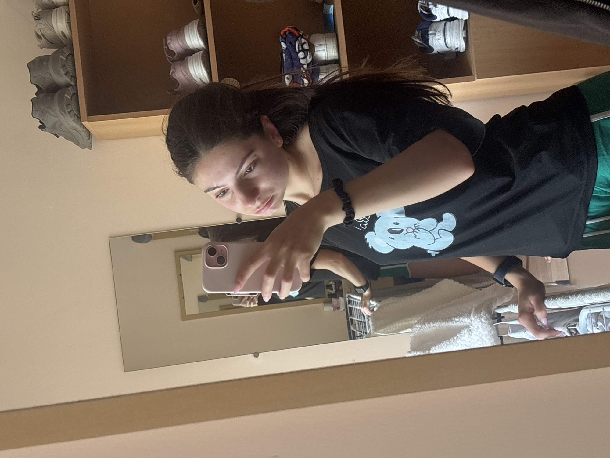 The image shows a young woman in a black t-shirt with a cartoon koala print, taking a selfie in a mirror. She has dark hair pulled back in a ponytail and is wearing a black scrunchie bracelet. She is looking at the camera with a neutral expression.
The background consists of shelves filled with shoes and other items. The shelves appear to be made of light-colored wood. In the foreground, a mirror reflects the woman and her surroundings. The overall lighting suggests it is daytime, and the setting appears to be a room, possibly a bedroom or dressing area, given the presence of shoes and what looks like a shelf for clothing. The image is taken indoors. There is no visible text in the image. The location context is Varna, Bulgaria.