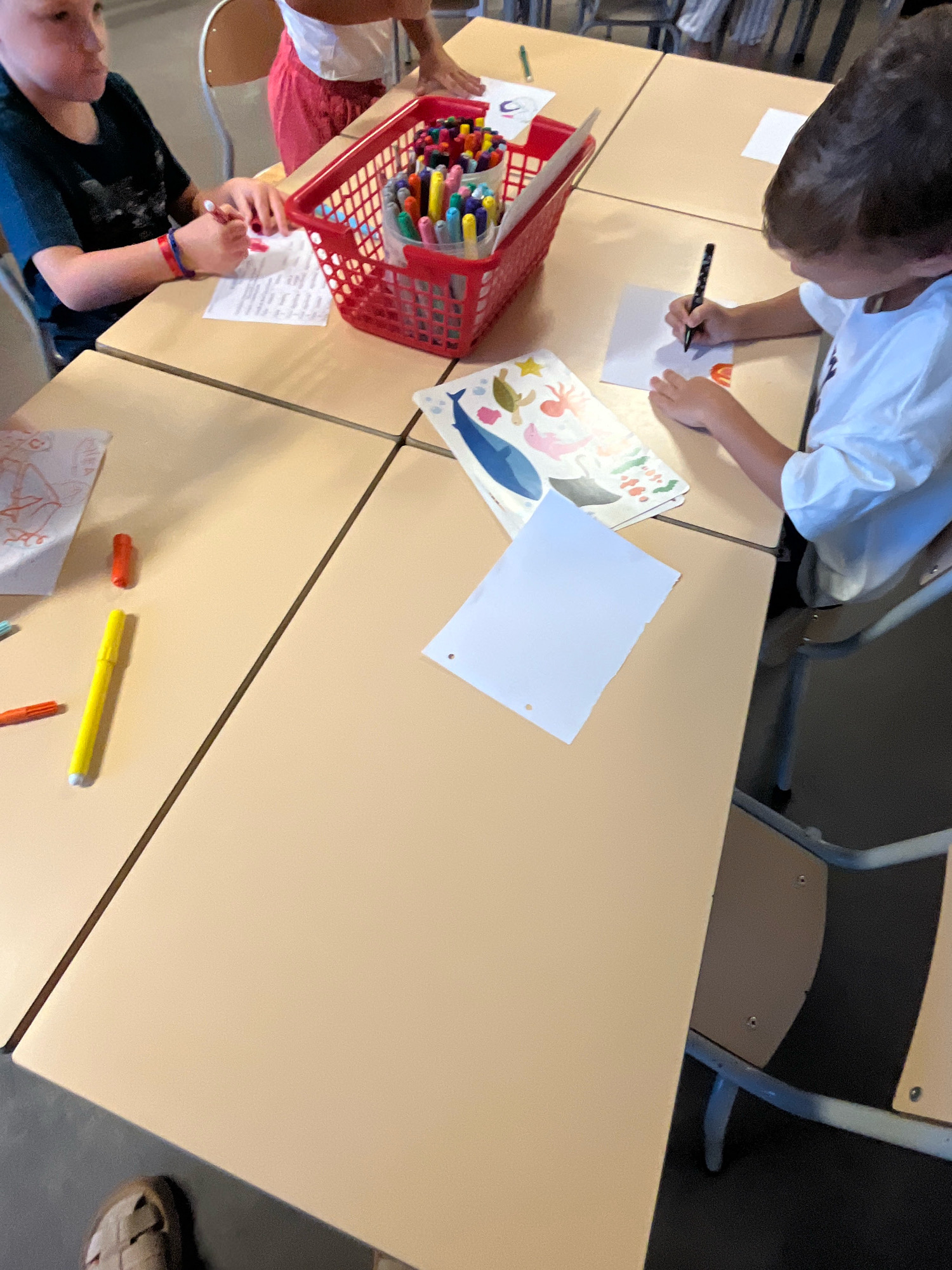 The image depicts a close-up, eye-level view of children engaged in drawing and coloring activities at a table. The scene appears to be in a classroom or community center setting in La Fouillouse, France, as indicated by the presence of multiple tables and chairs designed for group activities.
In the foreground, a child wearing a dark t-shirt is drawing on a piece of paper, holding a red marker. They have a red and blue bracelet on their left wrist. To their right is a red plastic basket overflowing with colorful markers and pens.
A second child, wearing a white t-shirt, is drawing on a small square of paper with a black marker. Their left hand is resting on the paper, stabilizing it.
Scattered on the table are various art supplies, including individual markers, a yellow highlighter, and sheets of paper with drawings. One drawing on the left features a red outline of a figure, and another in the center shows a colorful depiction of marine life, including a whale, a turtle, and coral.
The table itself is composed of four light-colored, rectangular sections, suggesting a modular table setup. In the background, a third person, partially visible, appears to be a child wearing a white top and red shorts, also engaged in an activity. The environment is lit with bright, natural light. The image provides a snapshot of a creative and educational moment.