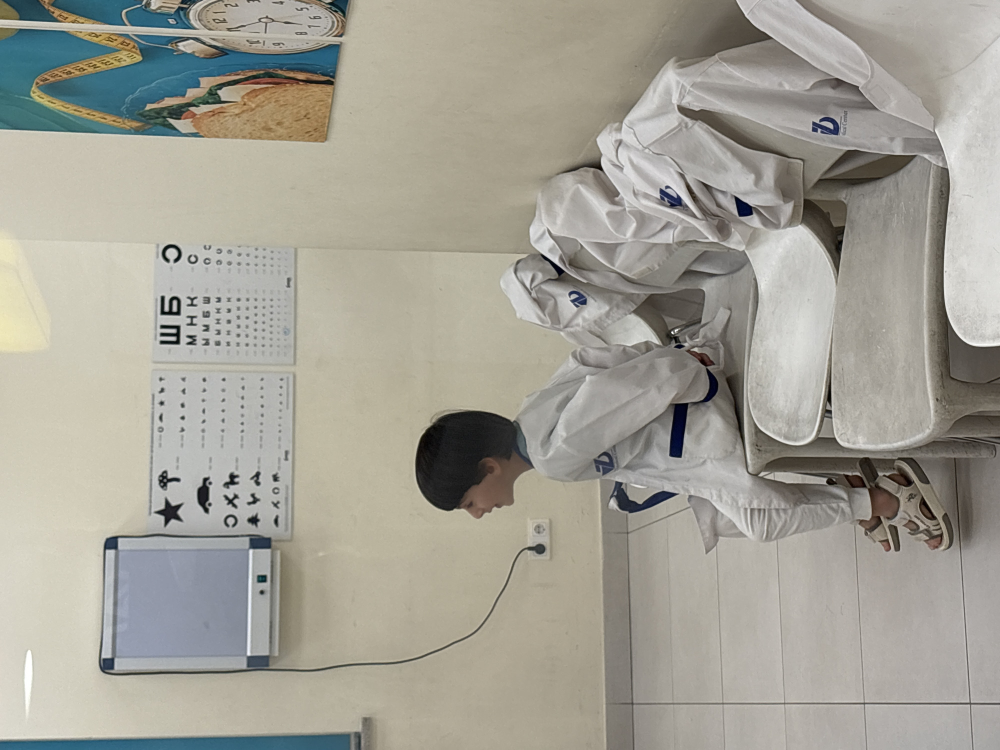 This is a vertically oriented, eye-level, medium shot of a young boy wearing a white karate uniform with blue trim. He is standing in what appears to be a medical clinic or examination room. The boy is facing left and looking down, with his arms crossed in front of him. He has short, dark hair.
Behind the boy on the wall are two eye charts, one with letters and numbers, and another with simple shapes like a star, a mushroom, a silhouette of a person, and an animal. Below the charts is a light box, typically used for viewing X-rays. A black electrical cord runs from an outlet on the wall to the light box.
To the right of the boy, a stack of white lab coats is draped over a white plastic chair. The lab coats have a blue logo that reads "Dical Center". The floor is tiled with white squares.
In the upper left corner of the image, a poster is partially visible. It features a clock face and a sandwich with a measuring tape wrapped around it, suggesting a theme of health or fitness.
The scene suggests a moment of waiting or perhaps a brief break during an appointment. The lighting is bright and consistent, indicating an indoor setting, likely during daylight hours. The boy's posture and downward gaze could indicate thoughtfulness, shyness, or perhaps mild boredom. The presence of medical equipment and eye charts firmly places the scene in a clinical environment. The location context of Yerevan, Armenia, is noted, but no specific visual cues within the image directly confirm this.