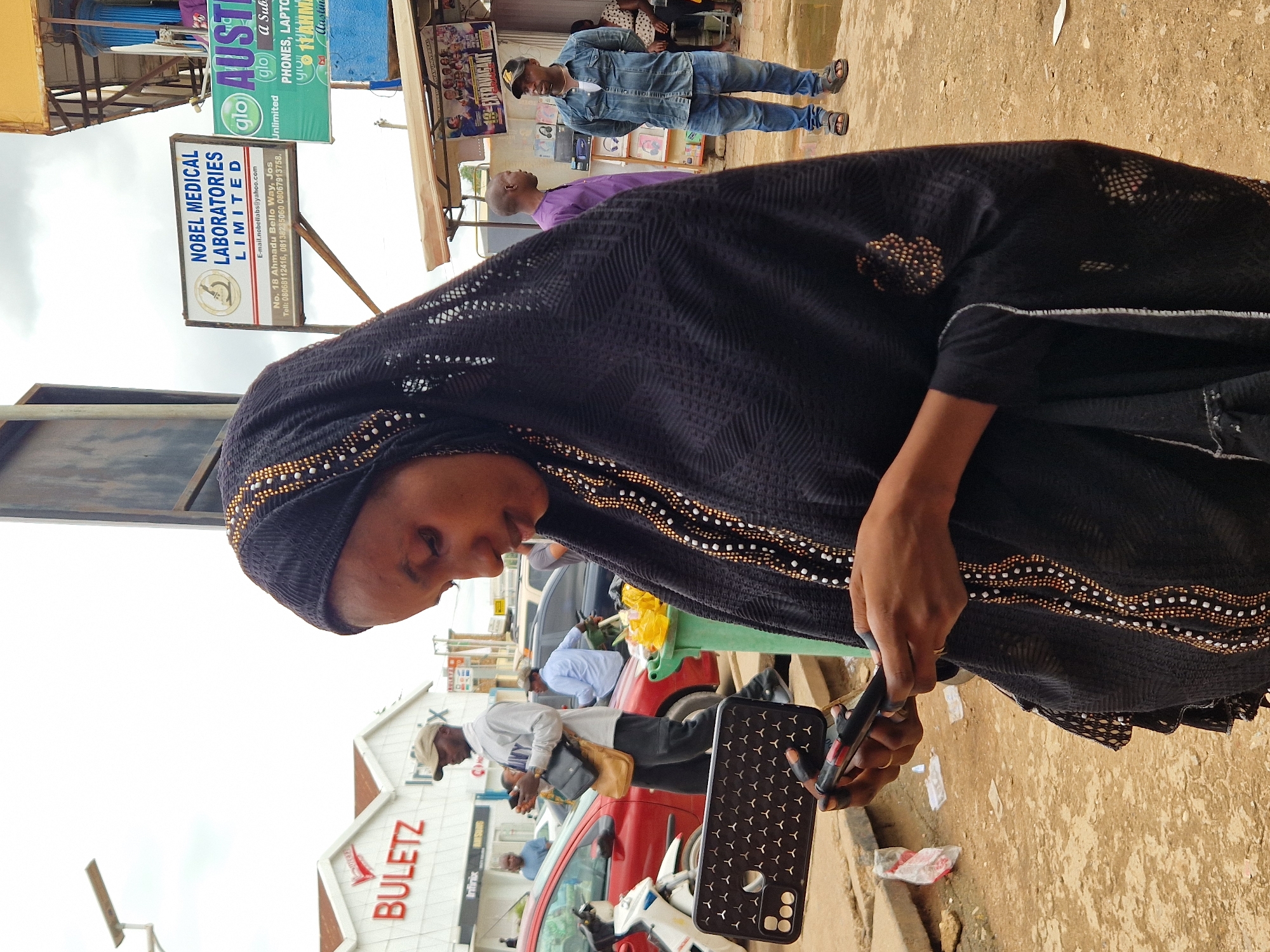 This image depicts a street scene with several people and commercial establishments. In the foreground, a woman wearing a black hijab and traditional Islamic attire is looking down at her phone. She has decorative beads and patterns on her garment. Her right hand is holding a phone case, and she appears to be using or examining a phone.
In the background, there are multiple signs for businesses. One prominent sign reads "BULETZ" in large red letters, suggesting a retail store. Another sign indicates "NOBEL MEDICAL LABORATORIES LIMITED" with contact information. There are also signs for "glo" and "Austin," possibly related to telecommunications or retail services. The presence of various signs and people suggests a bustling commercial area.
Several individuals are visible in the midground and background, including men in casual clothing and a man wearing a blue denim jacket and jeans. Some people appear to be walking or standing around, engaged in their daily activities. There are also vehicles, including a red car, indicating a street or road setting.
The overall scene appears to be outdoors during daylight, though the exact time of day is not precisely discernible. The weather seems to be overcast, given the lighting conditions. The environment looks like a typical urban street in an area where outdoor commerce is common. The activity is focused on daily commerce and personal engagement with mobile devices.